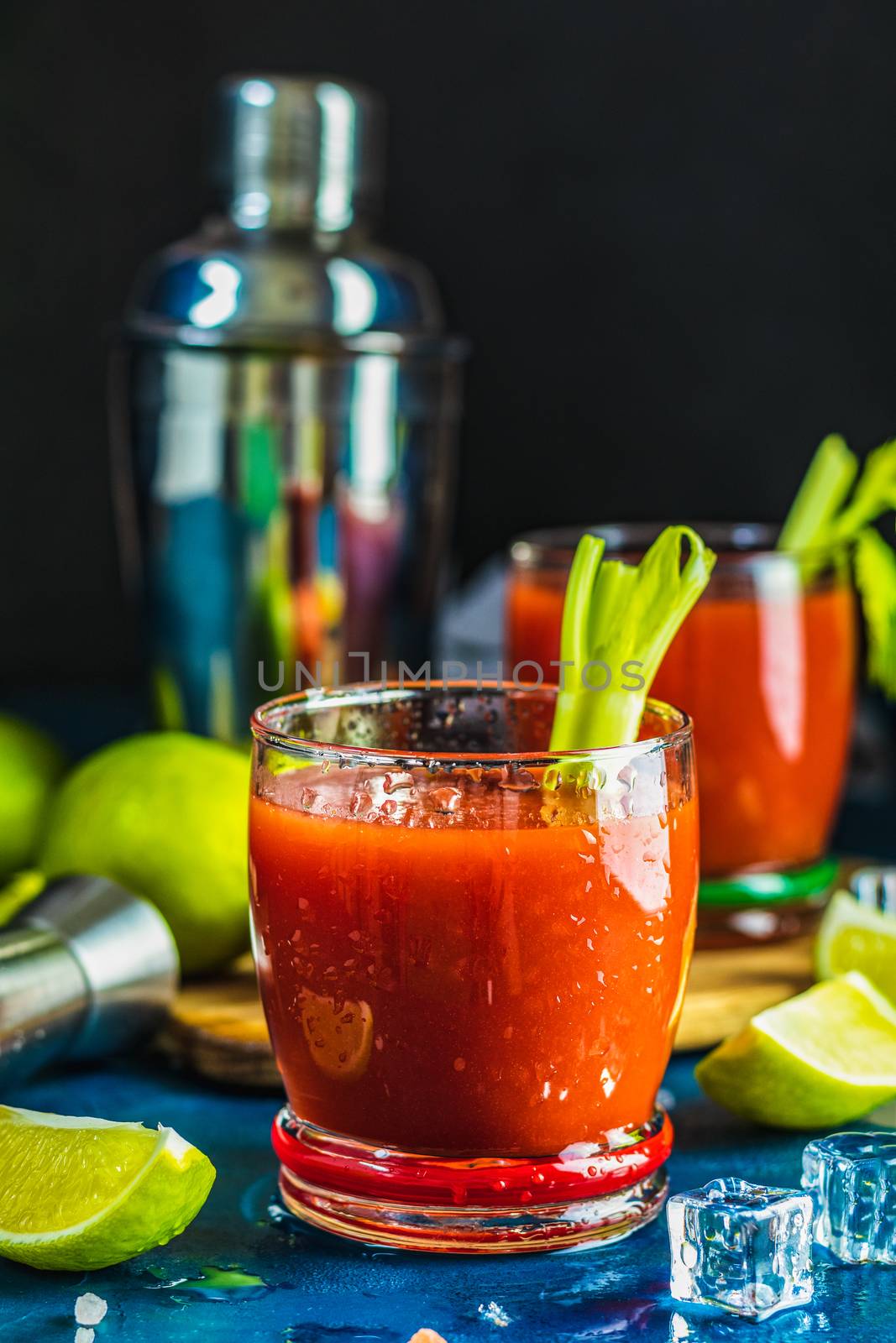 Tomato juice with celery, spices, salt and ice in portion drink glasses with copy space. Bloody Mary cocktail. Alcoholic drink and ingredients at dark blue concrete table surface.