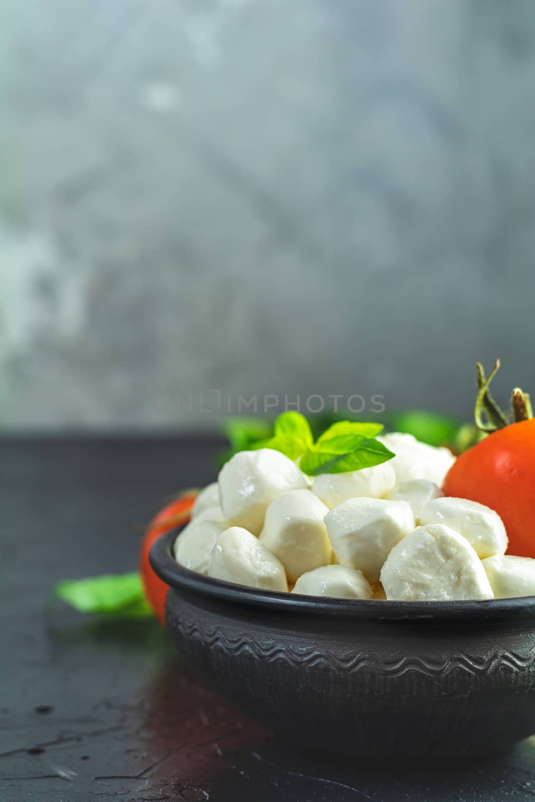 Mozzarella balls, buffalo in black ceramic plate, tomatoes and basil over dark background. With space. Rustic style. Ingredients for italian caprese salad. 