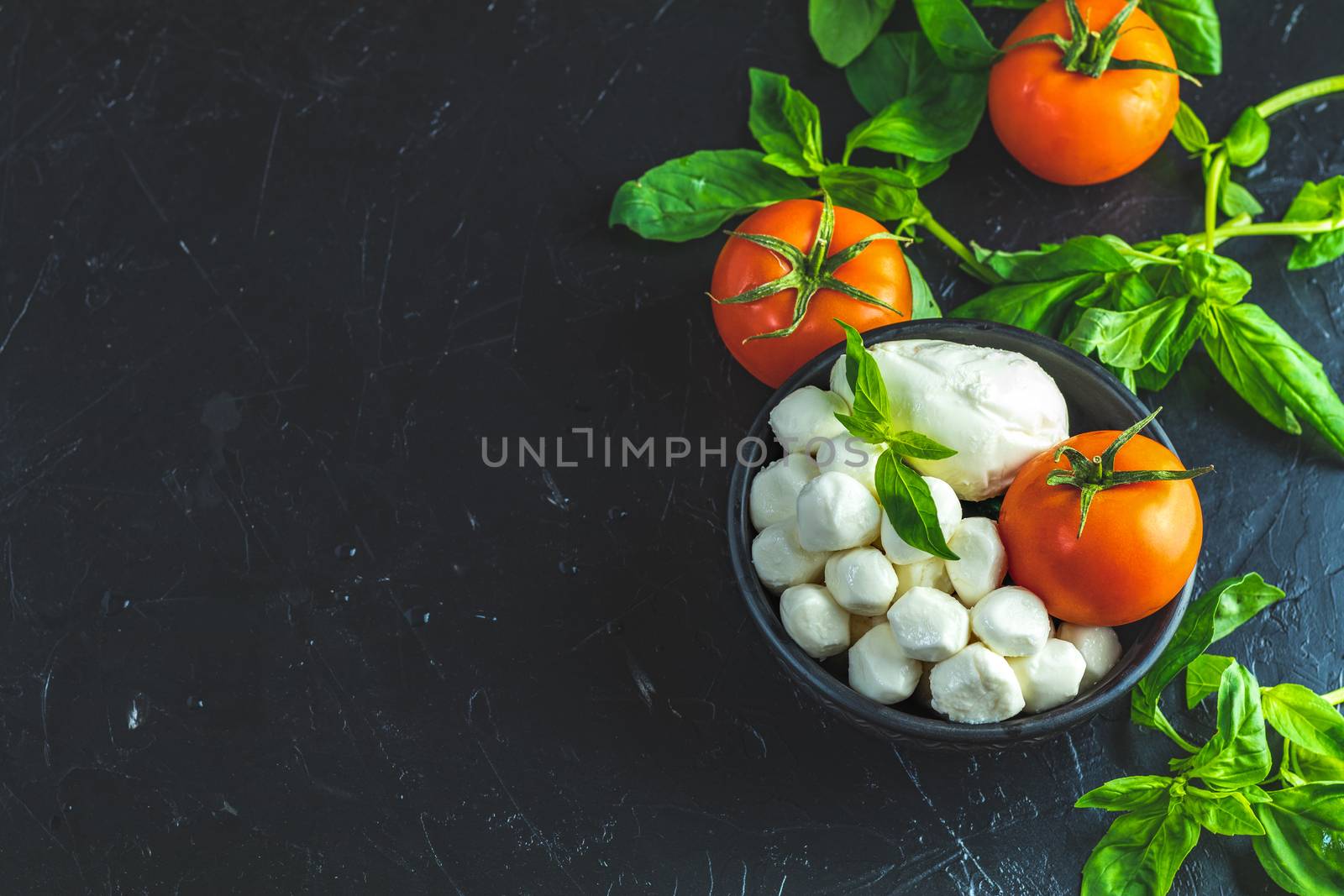 Mozzarella balls, buffalo in black ceramic plate, tomatoes and basil over dark background. With space. Rustic style. Ingredients for italian caprese salad