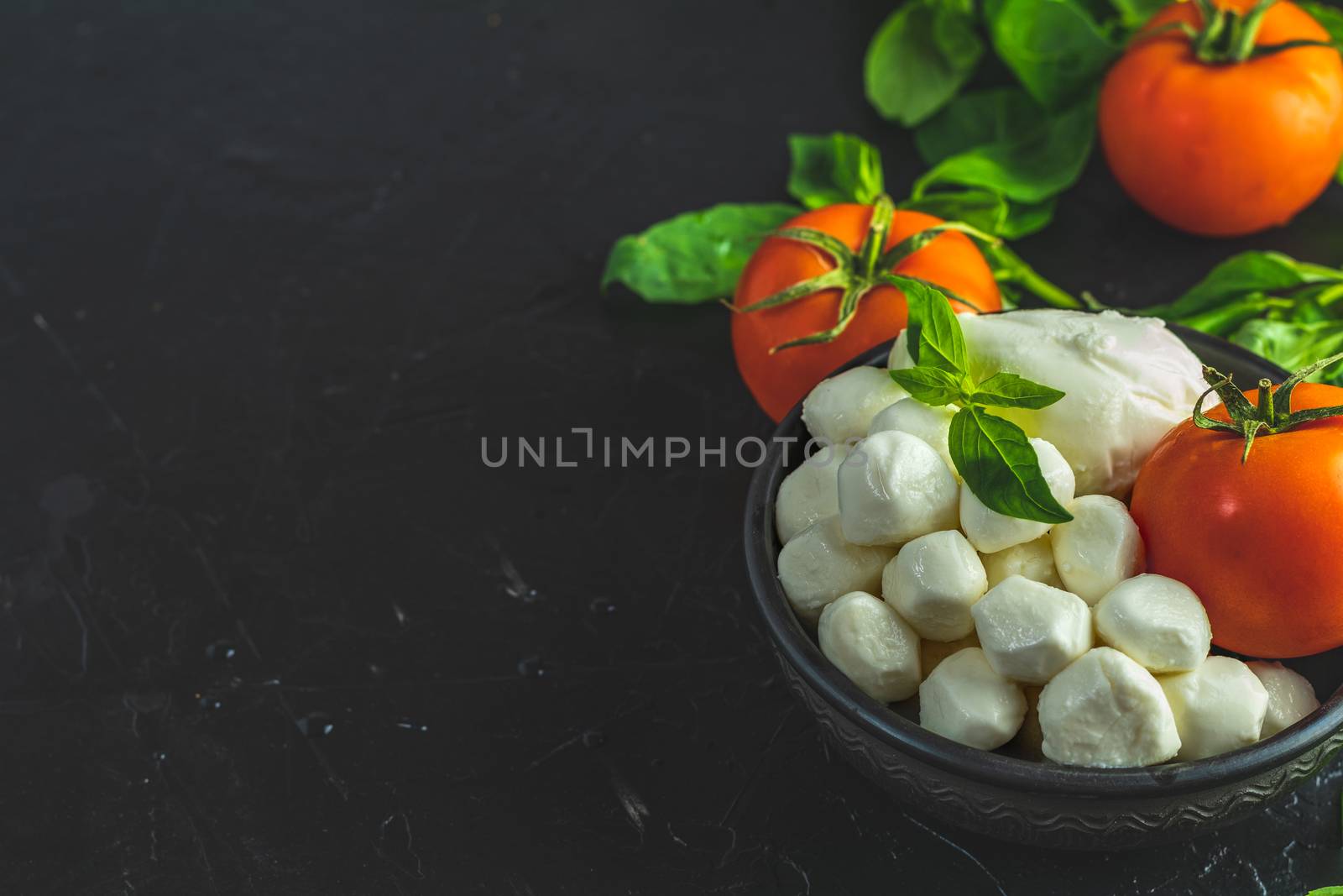 Mozzarella balls, buffalo in black ceramic plate, tomatoes and basil over dark background. With space. Rustic style. Ingredients for italian caprese salad