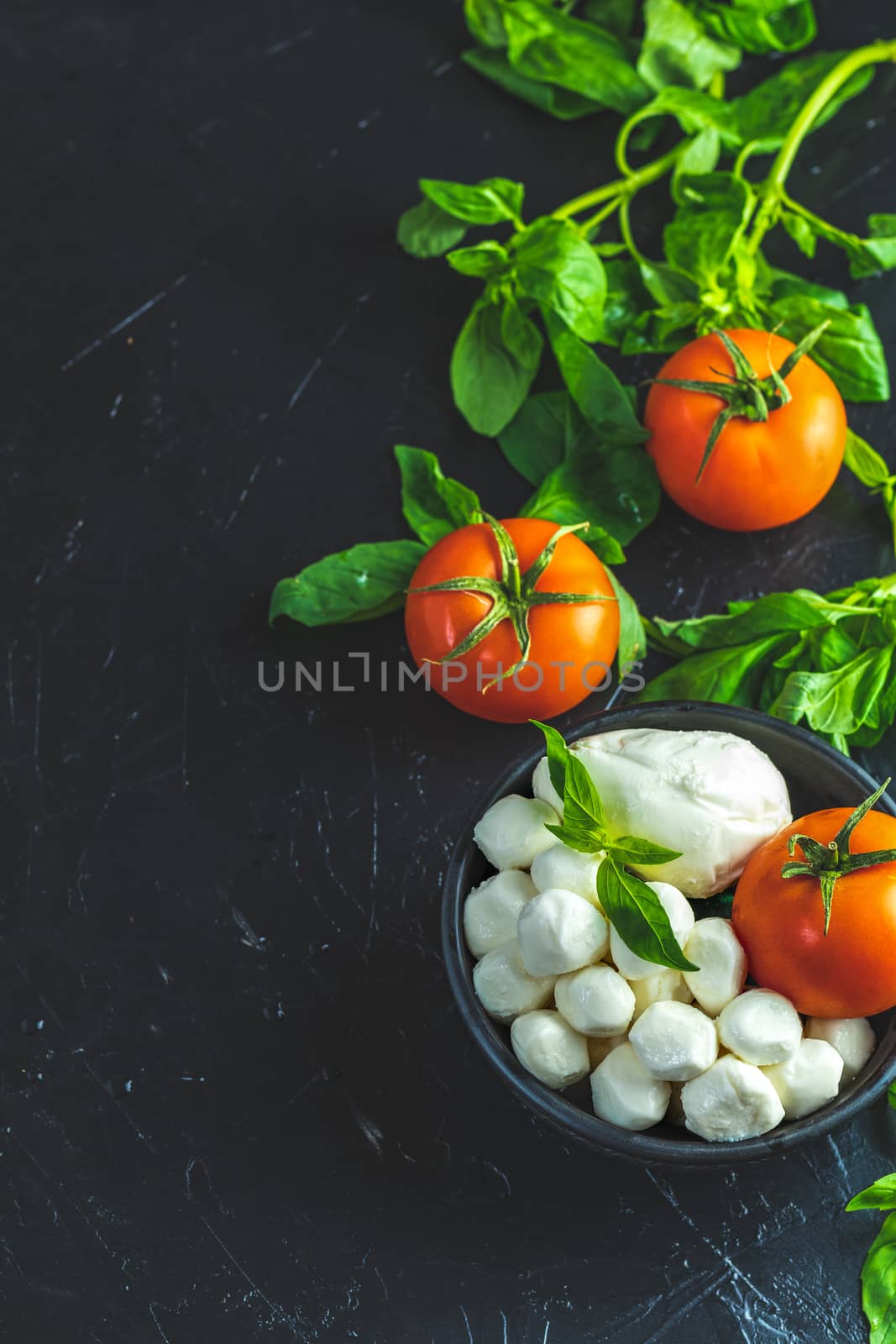 Mozzarella balls, buffalo in black ceramic plate, tomatoes and basil over dark background. With space. Rustic style. Ingredients for italian caprese salad. 