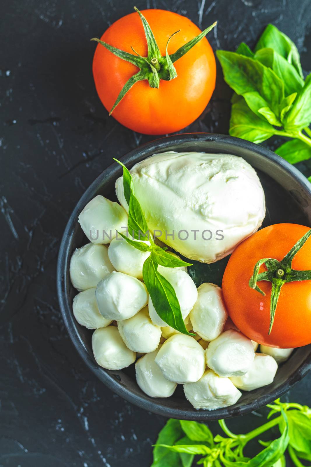 Mozzarella balls, buffalo in black ceramic plate, tomatoes and basil over dark background. With space. Rustic style. Ingredients for italian caprese salad