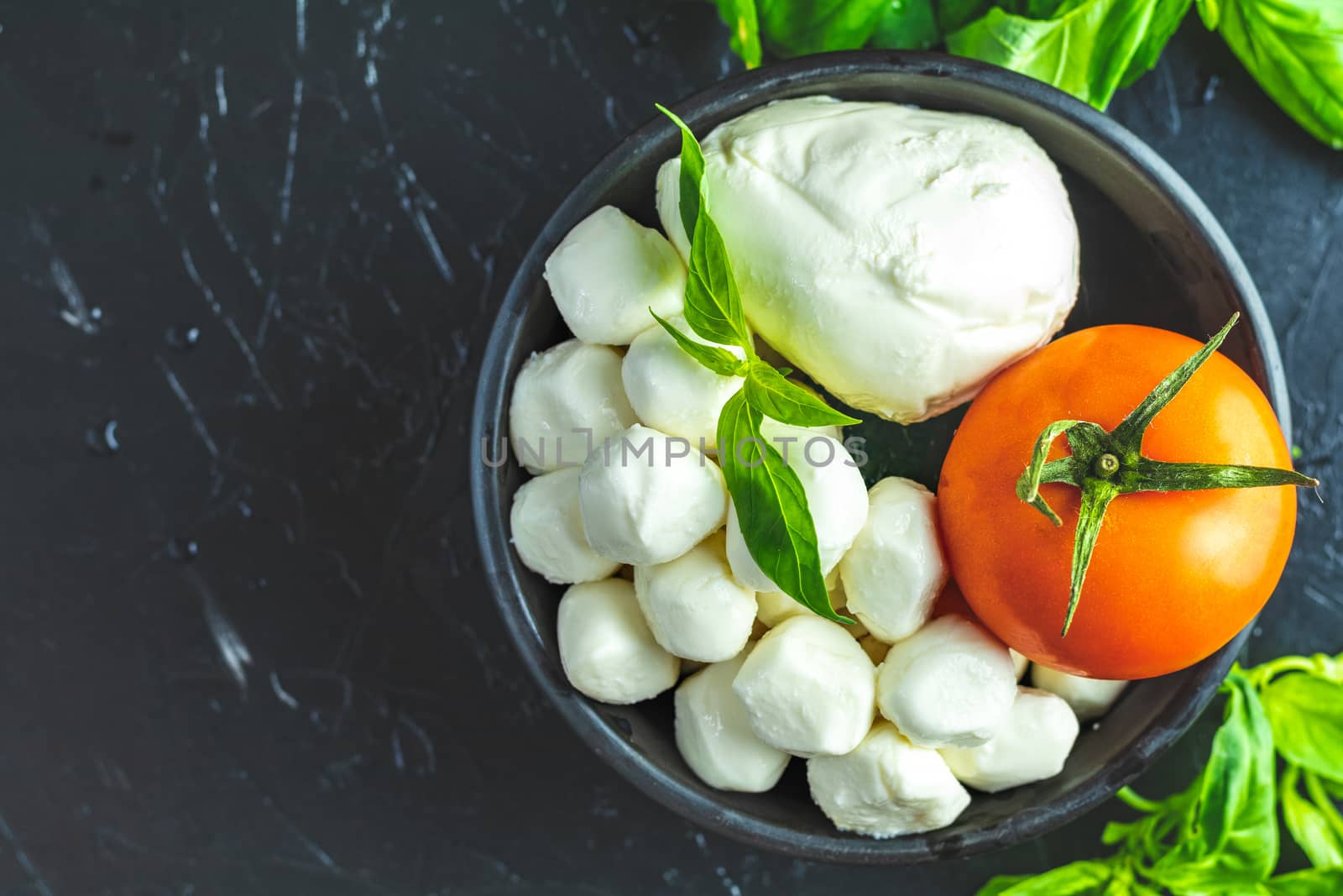Mozzarella balls, buffalo in black ceramic plate, tomatoes and basil over dark background. With space. Rustic style. Ingredients for italian caprese salad. 