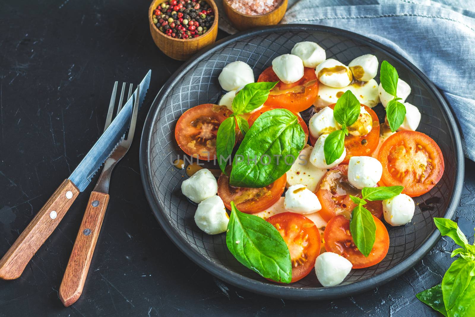 Italian caprese salad with sliced tomatoes, mozzarella cheese, basil and olive oil served in black ceramic plate with fork and knife on textile napkin over dark concrete table surface with copy space.