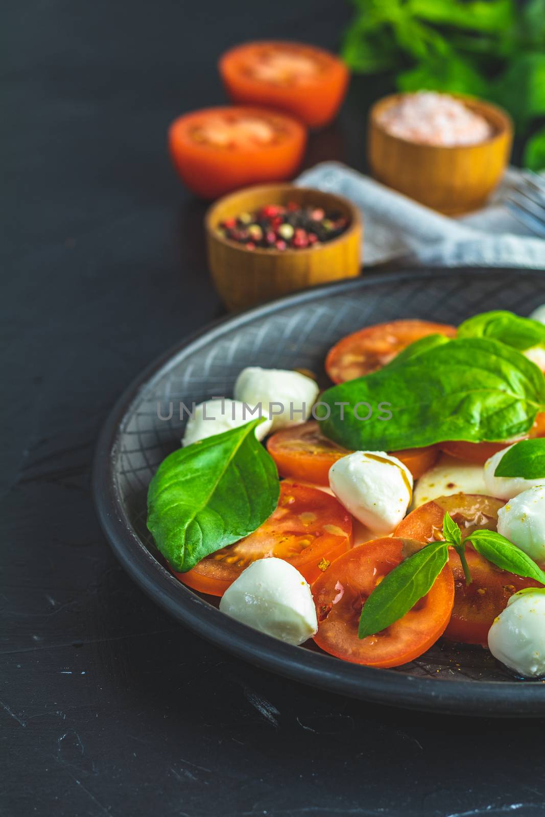 Italian caprese salad with sliced tomatoes, mozzarella cheese, basil and olive oil served in black ceramic plate with fork and knife on textile napkin over dark concrete table surface with copy space