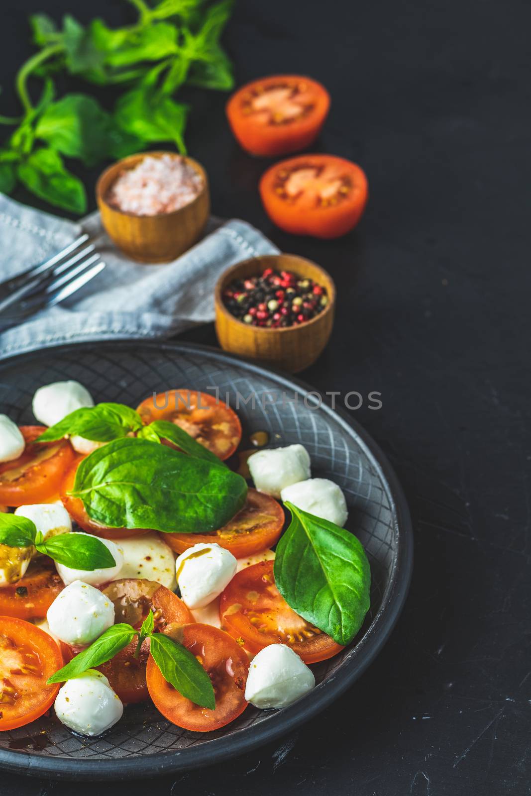 Italian caprese salad with sliced tomatoes, mozzarella cheese, basil and olive oil served in black ceramic plate with fork and knife on textile napkin over dark concrete table surface with copy space.