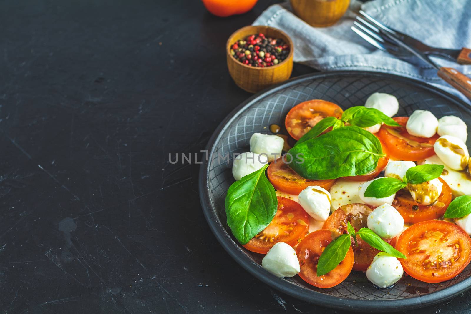 Italian caprese salad with sliced tomatoes, mozzarella cheese, basil and olive oil served in black ceramic plate with fork and knife on textile napkin over dark concrete table surface with copy space.