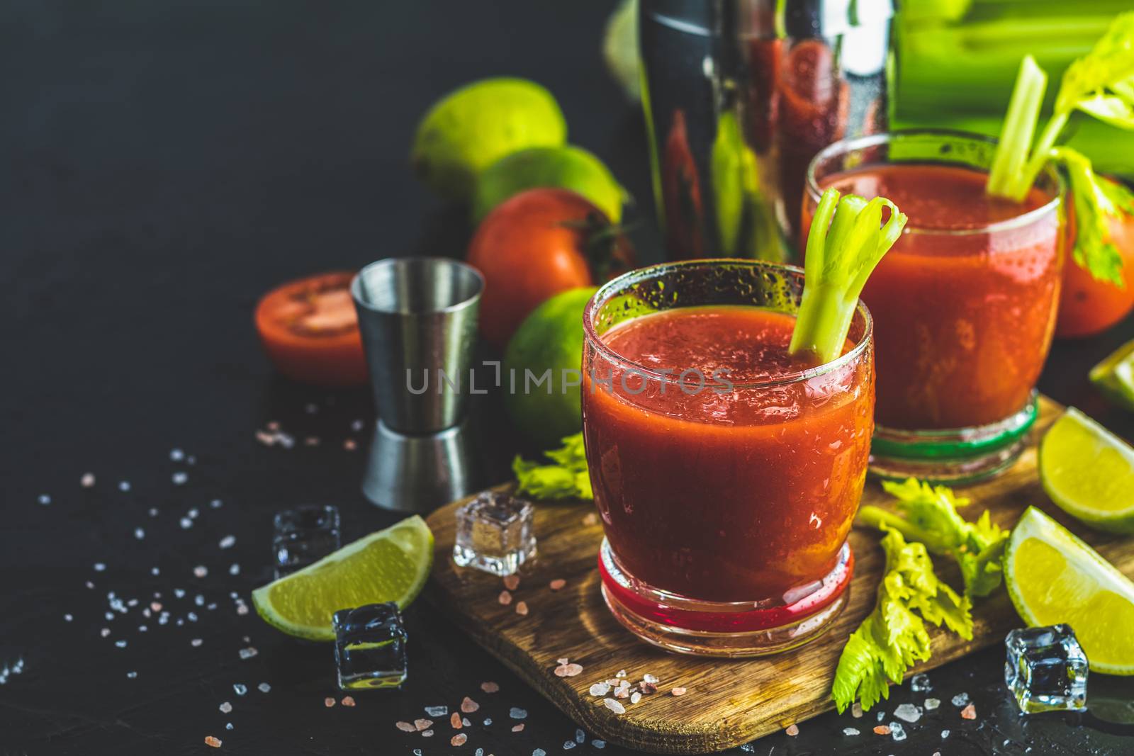 Tomato juice with celery, spices, salt and ice in portion glasses with copy space. Bloody Mary cocktail. Alcoholic drink and ingredients at dark concrete table surface.