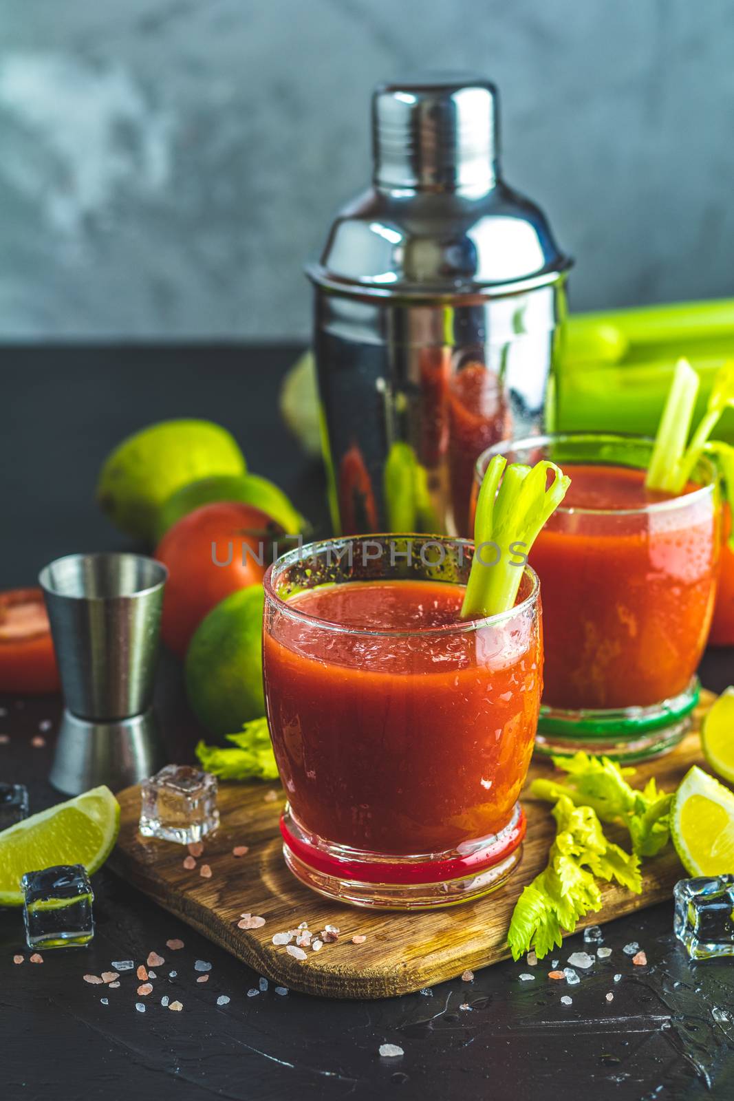Tomato juice with celery, spices, salt and ice in portion glasses with copy space. Bloody Mary cocktail. Alcoholic drink and ingredients at dark concrete table surface.