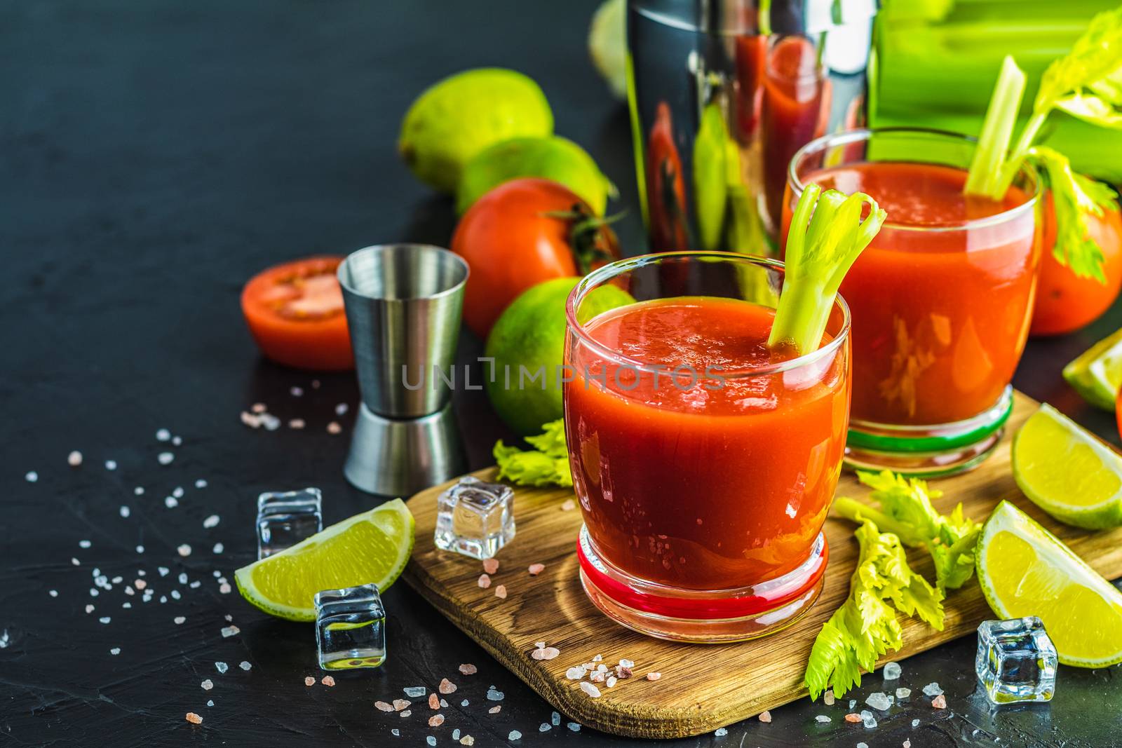 Tomato juice with celery, spices, salt and ice in portion glasses with copy space. Bloody Mary cocktail. Alcoholic drink and ingredients at dark concrete table surface. 