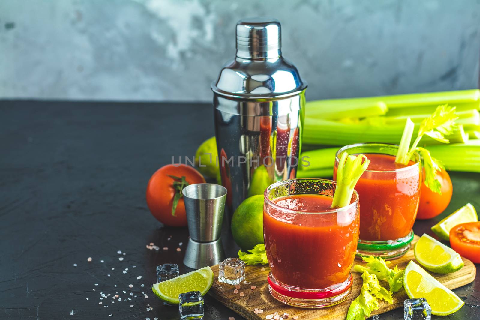 Tomato juice with celery, spices, salt and ice in portion glasses with copy space. Bloody Mary cocktail. Alcoholic drink and ingredients at dark concrete table surface. 