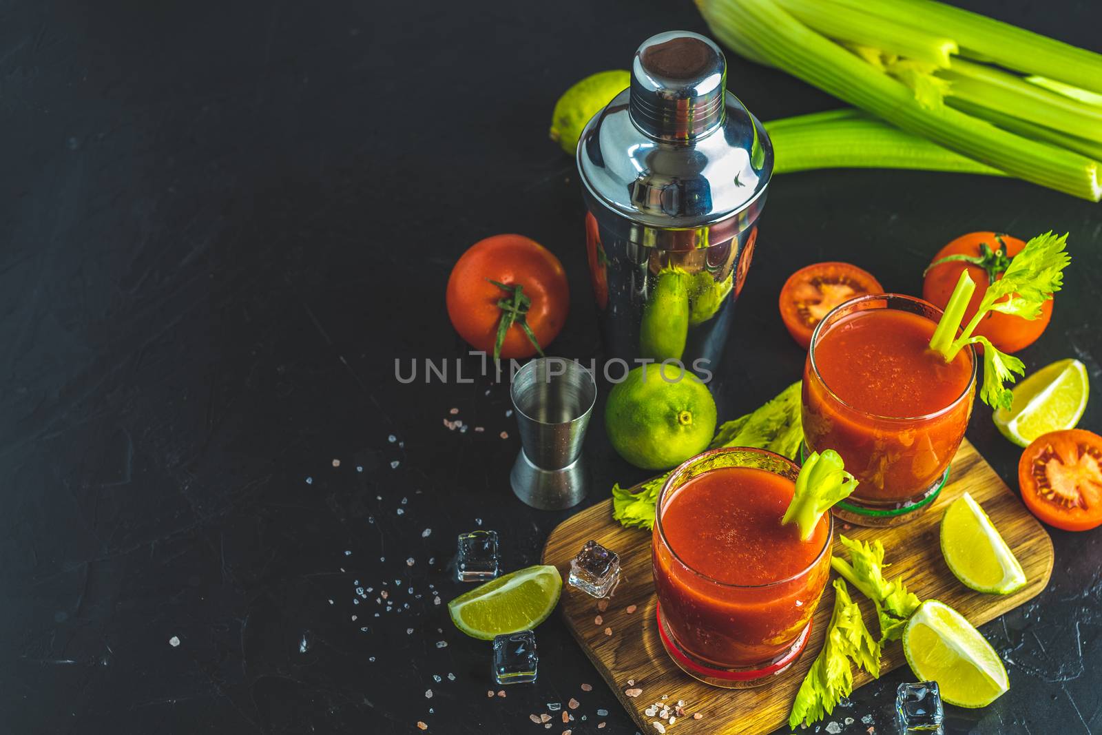 Tomato juice with celery, spices, salt and ice in portion glasses with copy space. Bloody Mary cocktail. Alcoholic drink and ingredients at dark concrete table surface.