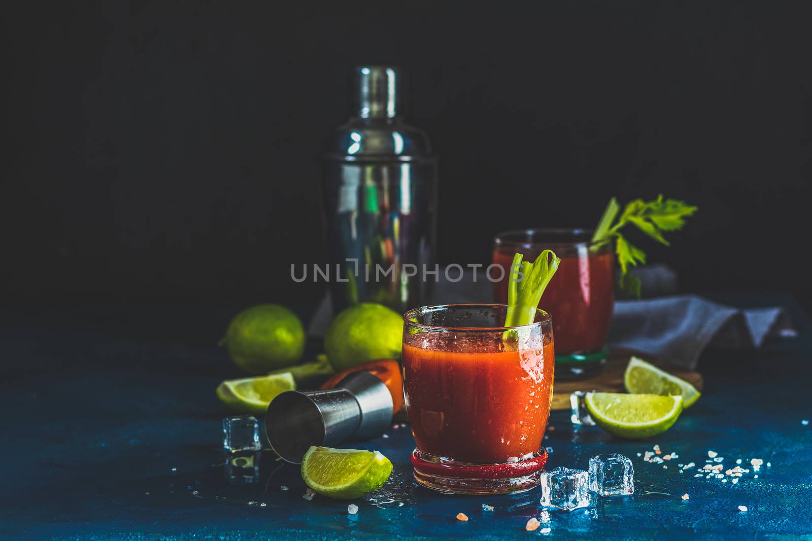 Tomato juice with celery, spices, salt and ice in portion drink glasses with copy space. Bloody Mary cocktail. Alcoholic drink and ingredients at dark blue concrete table surface.