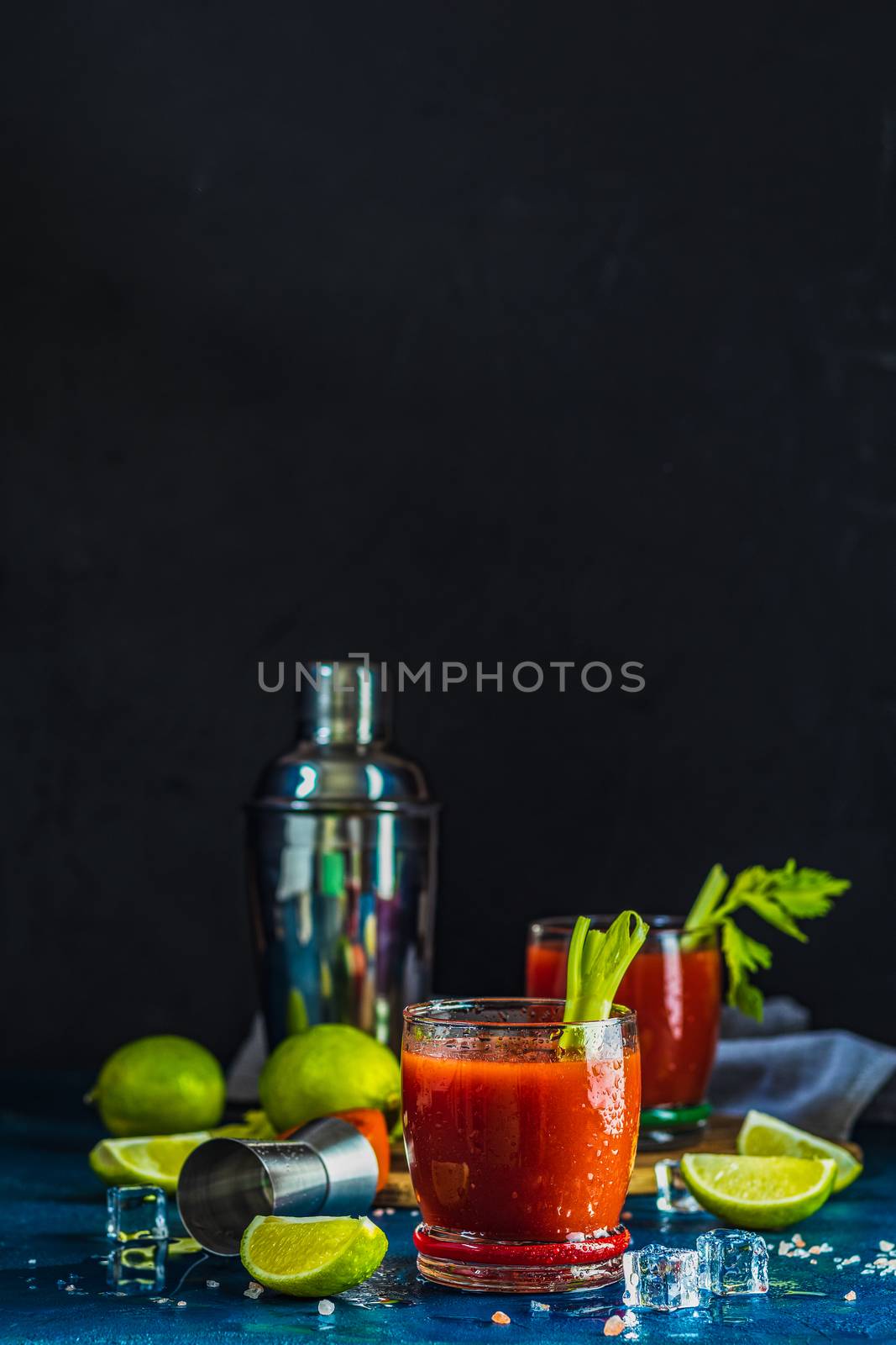 Tomato juice with celery, spices, salt and ice in portion drink glasses with copy space. Bloody Mary cocktail. Alcoholic drink and ingredients at dark blue concrete table surface.