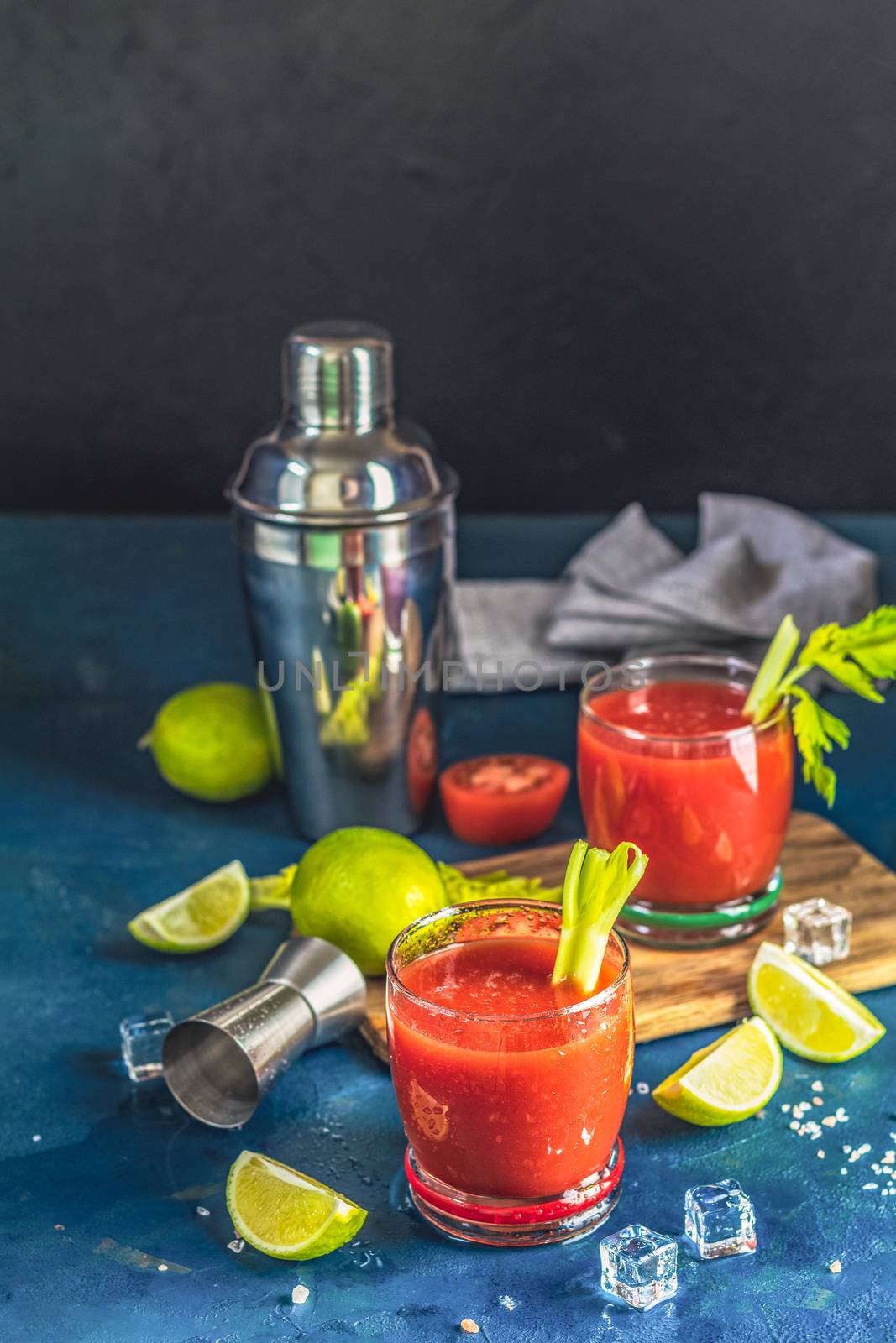 Tomato juice with celery, spices, salt and ice in portion drink glasses with copy space. Bloody Mary cocktail. Alcoholic drink and ingredients at dark blue concrete table surface.