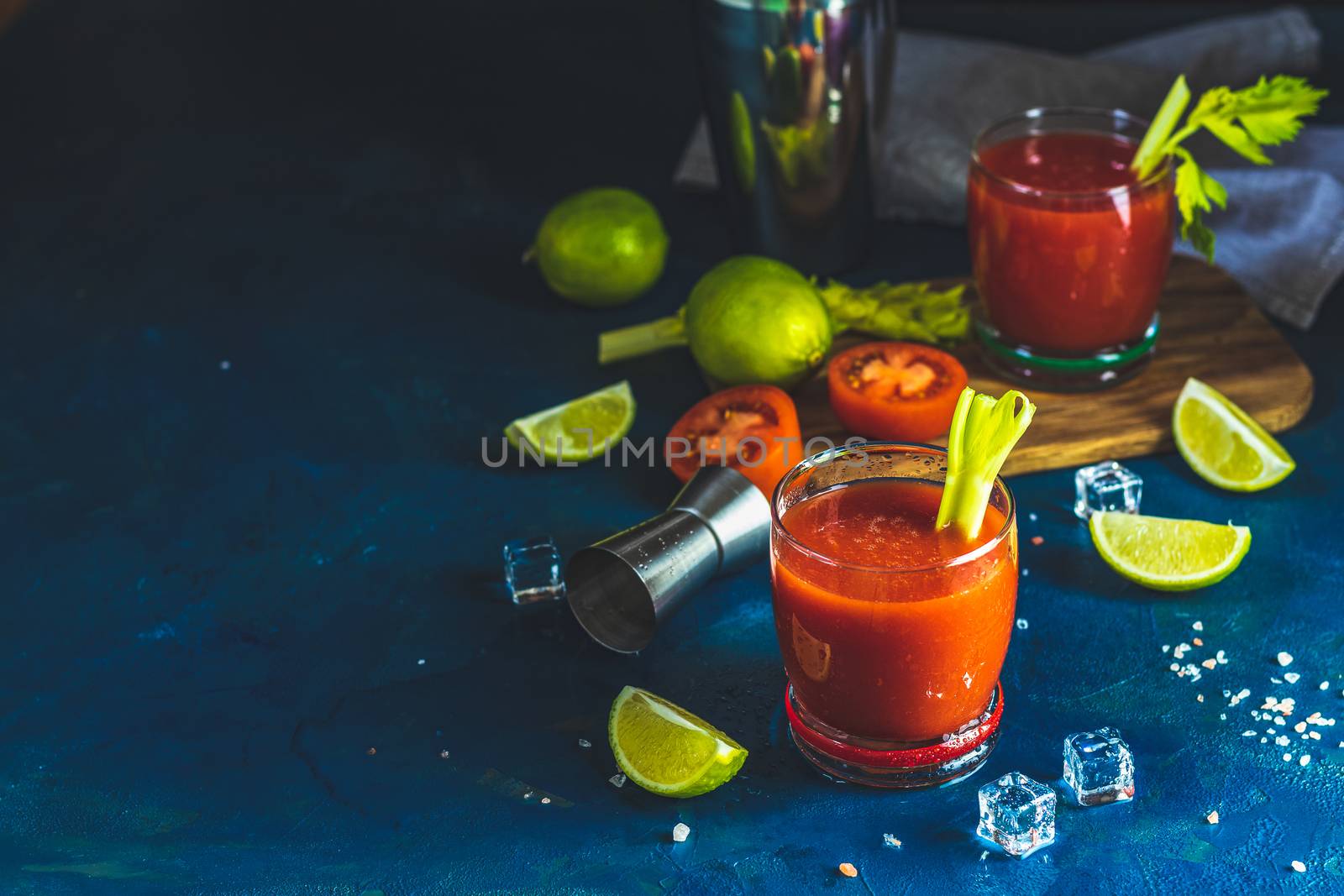Tomato juice with celery, spices, salt and ice in portion drink glasses with copy space. Bloody Mary cocktail. Alcoholic drink and ingredients at dark blue concrete table surface.