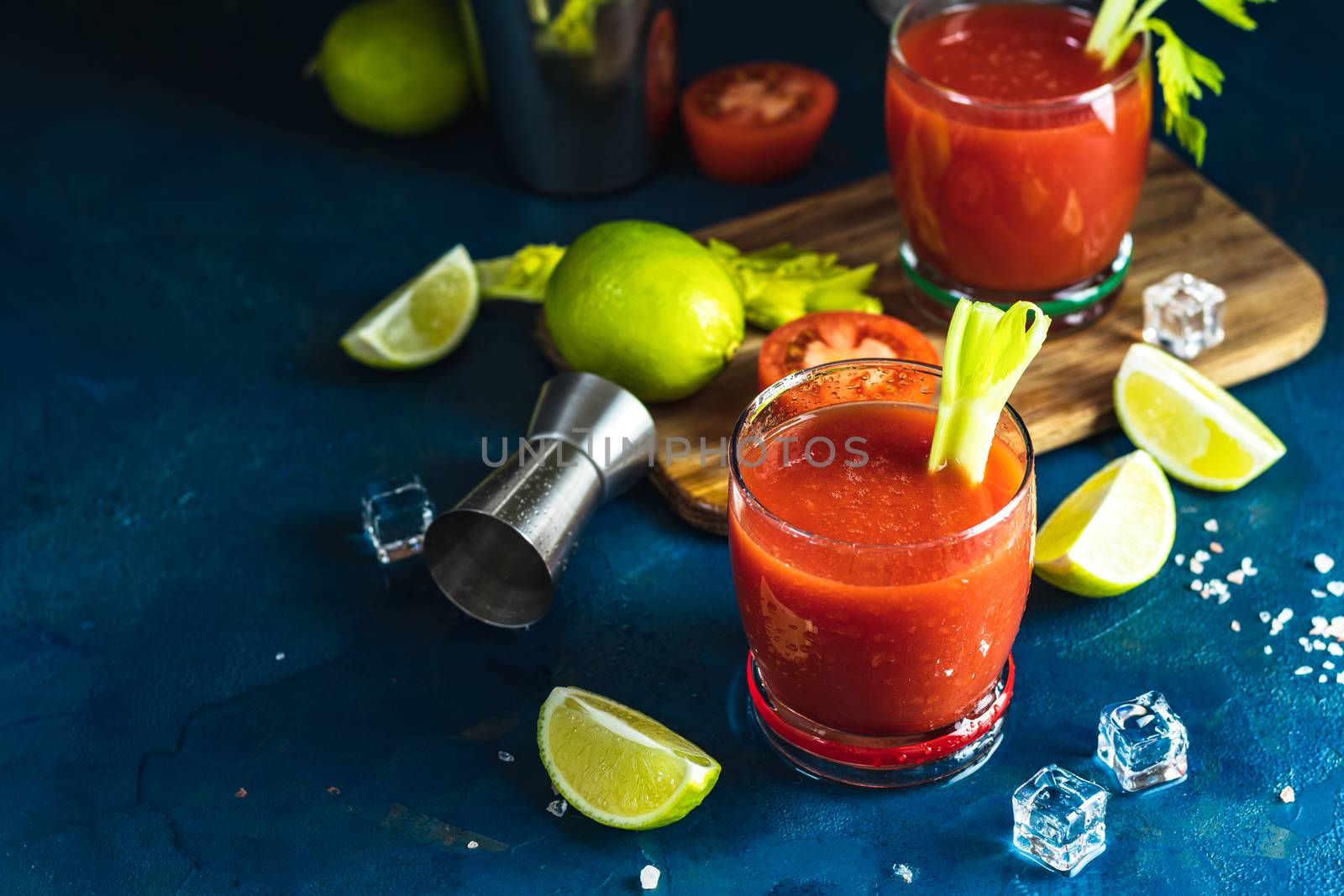 Tomato juice with celery, spices, salt and ice in portion drink glasses with copy space. Bloody Mary cocktail. Alcoholic drink and ingredients at dark blue concrete table surface.
