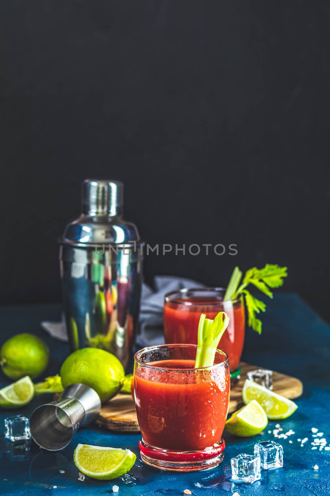 Tomato juice with celery, spices, salt and ice in portion drink glasses with copy space. Bloody Mary cocktail. Alcoholic drink and ingredients at dark blue concrete table surface.