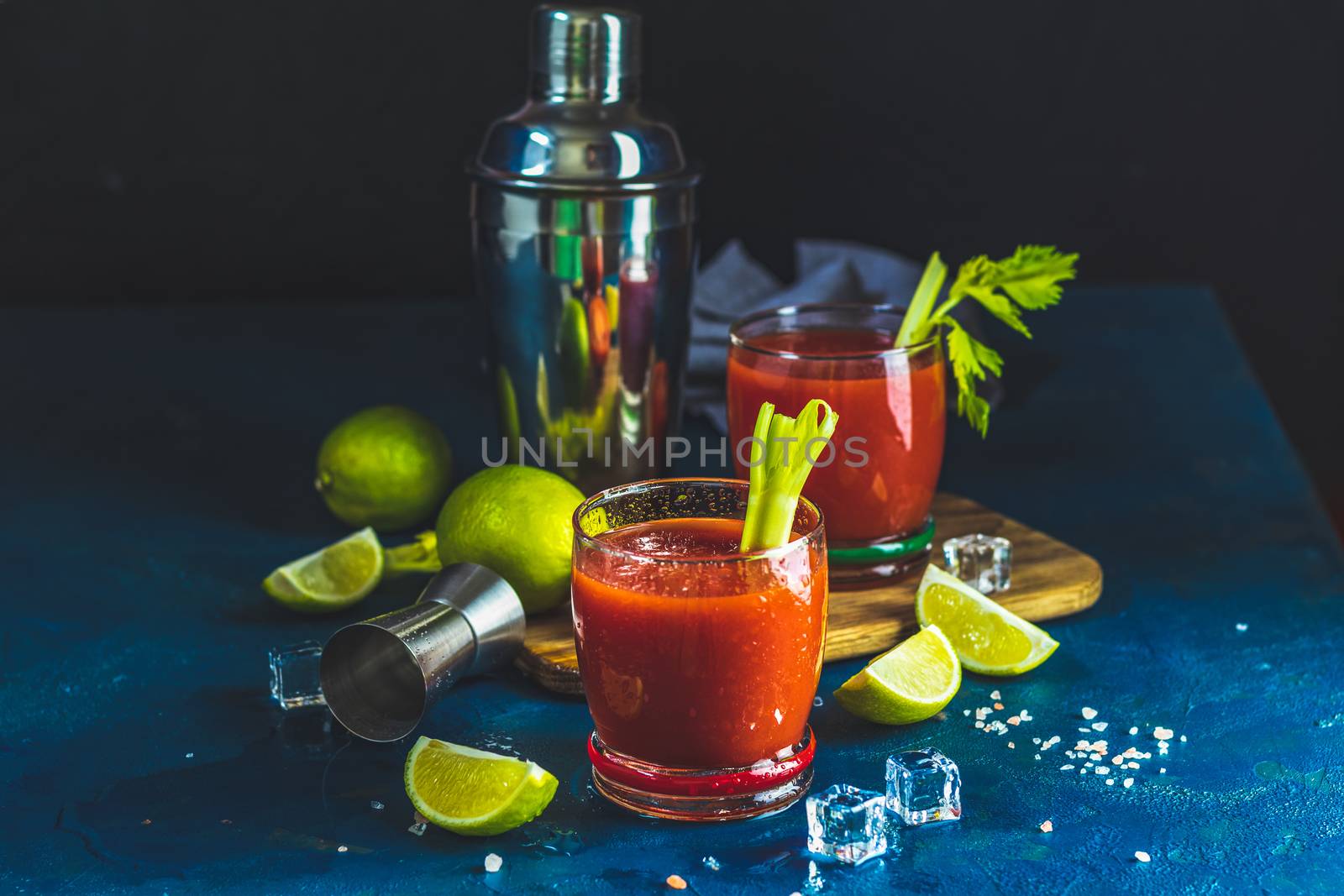 Tomato juice with celery, spices, salt and ice in portion drink glasses with copy space. Bloody Mary cocktail. Alcoholic drink and ingredients at dark blue concrete table surface.