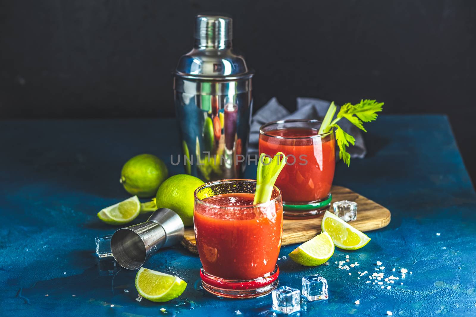 Tomato juice with celery, spices, salt and ice in portion drink glasses with copy space. Bloody Mary cocktail. Alcoholic drink and ingredients at dark blue concrete table surface.