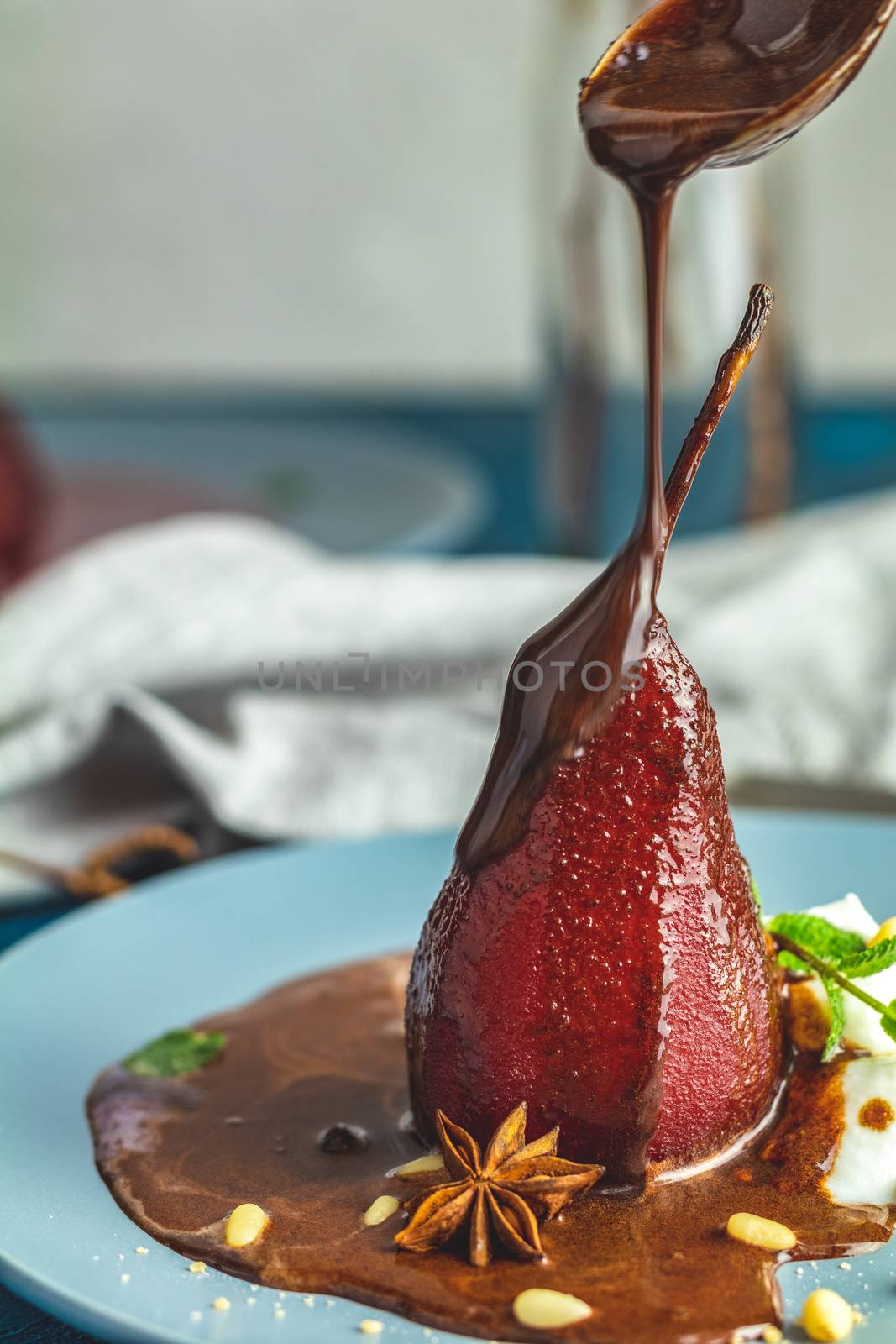 Chocolate sauce pours from a spoon on red pears in wine. Red pears are in a blue plate. The concept of still life on blue concrete surface.