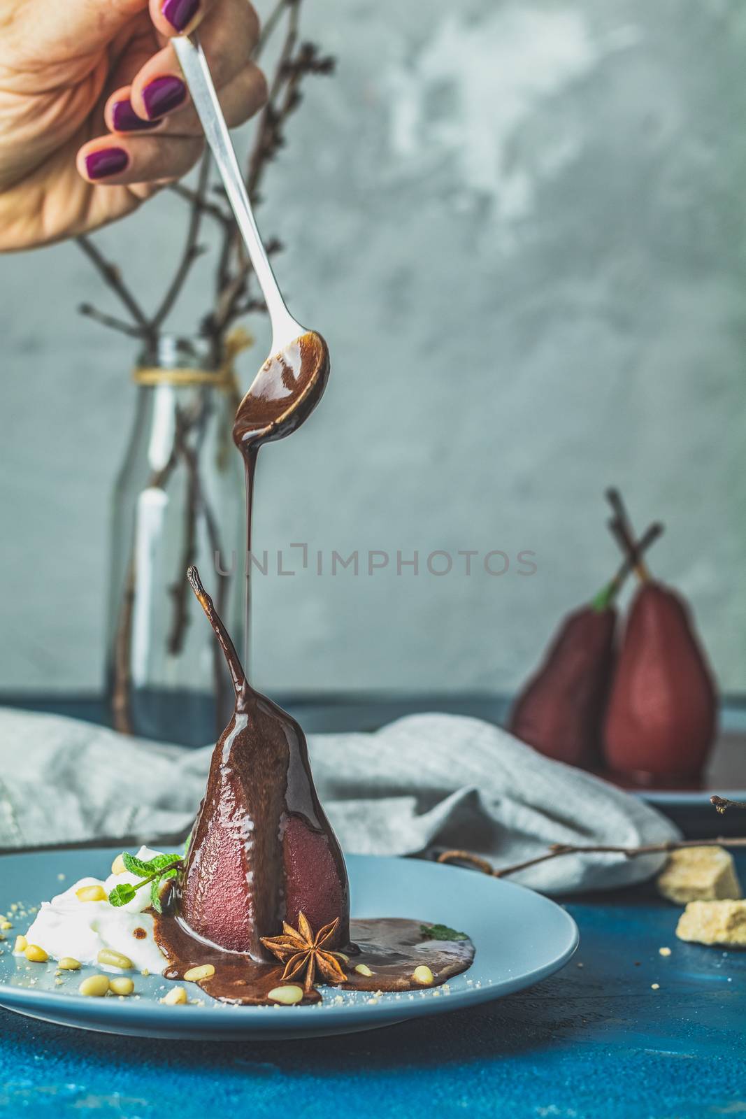 Chocolate sauce pours from a spoon on red pears in wine. Red pears are in a blue plate. The concept of still life on blue concrete surface