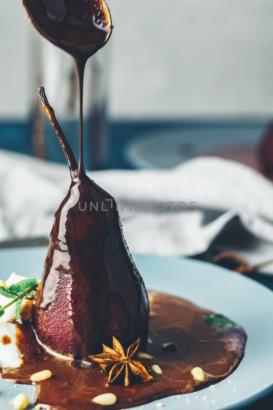 Chocolate sauce pours from a spoon on red pears in wine. Red pears are in a blue plate. The concept of still life on blue concrete surface.