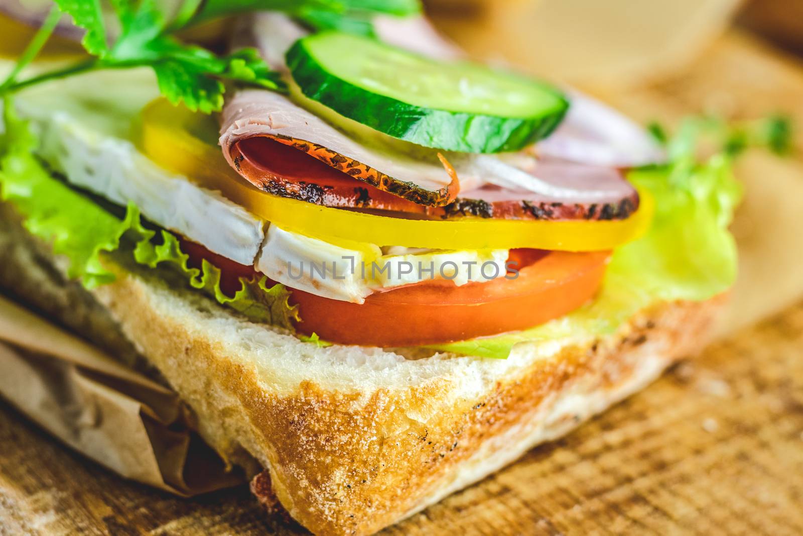 Ciabatta sandwich with ham, tomato, cheese, pepper, onion and salad on wooden cutting board with ingredients.