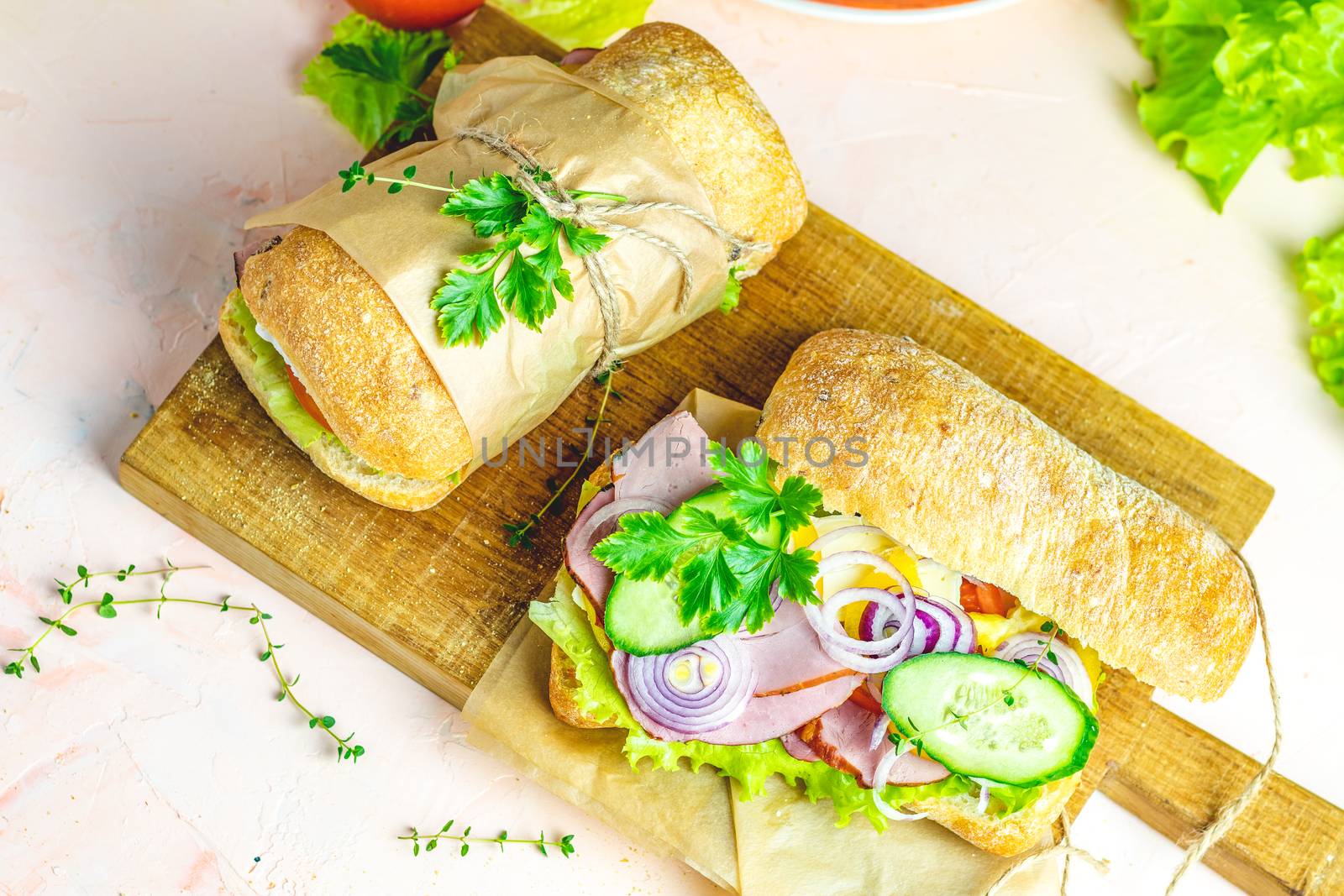 Ciabatta sandwich with ham, tomato, cheese, pepper, onion and salad on wooden cutting board with ingredients.