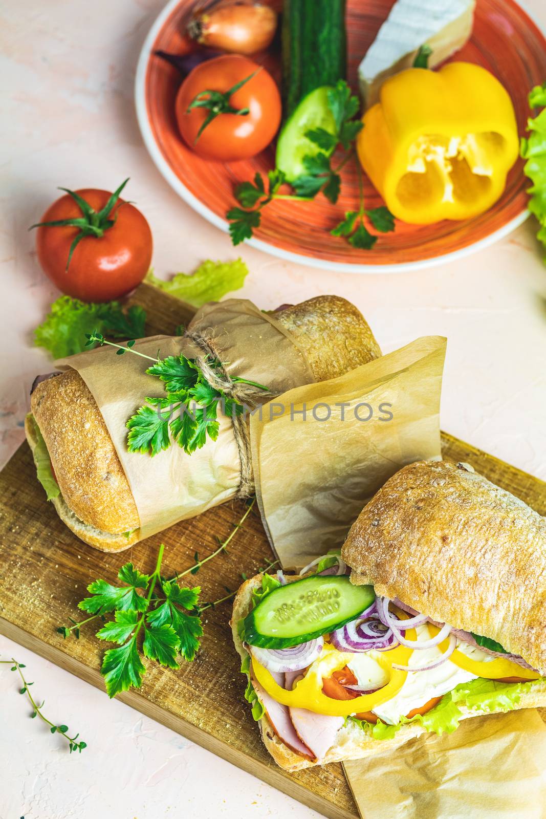 Ciabatta sandwich with ham, tomato, cheese, pepper, onion and salad on wooden cutting board with ingredients.