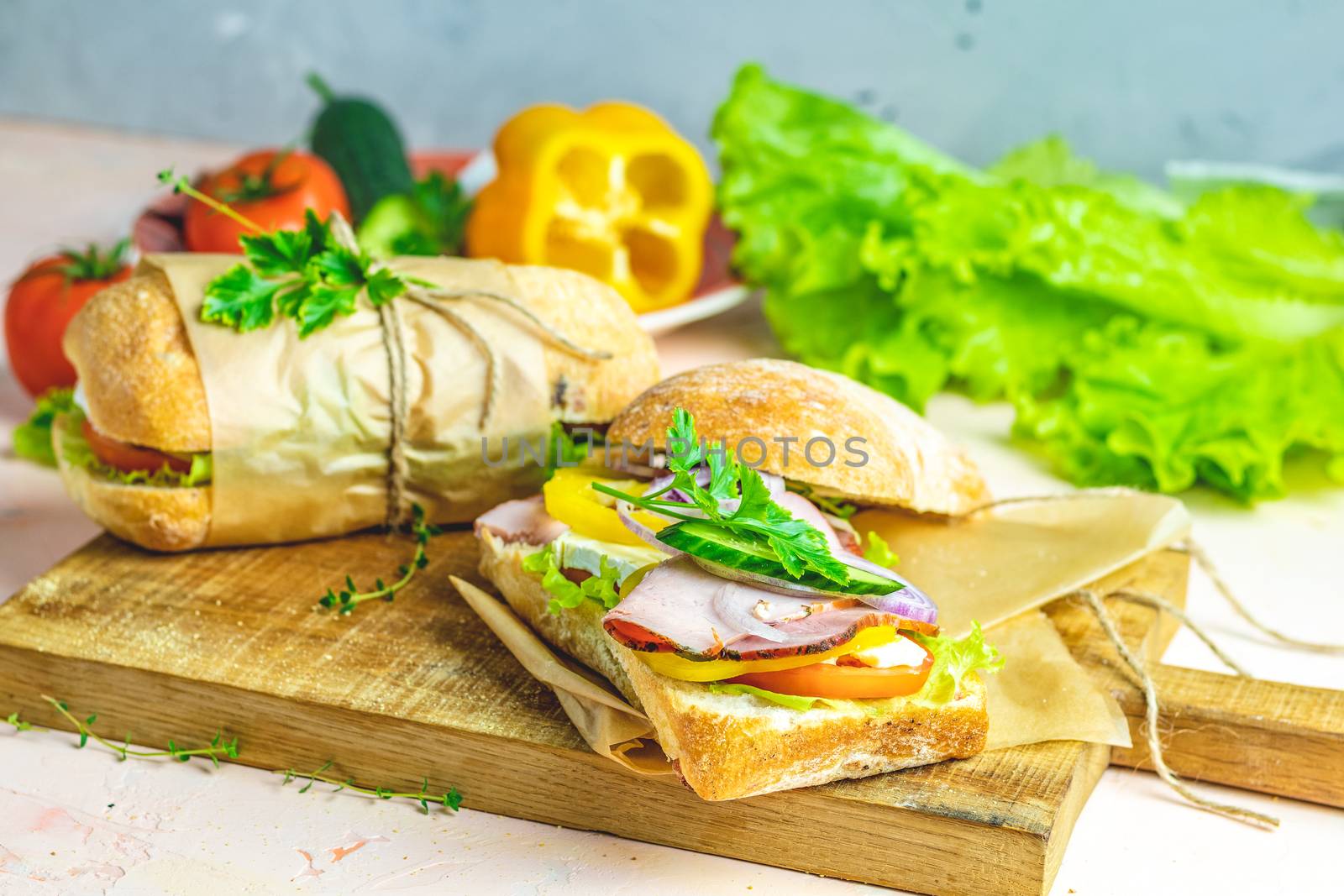 Ciabatta sandwich with ham, tomato, cheese, pepper, onion and salad on wooden cutting board with ingredients.