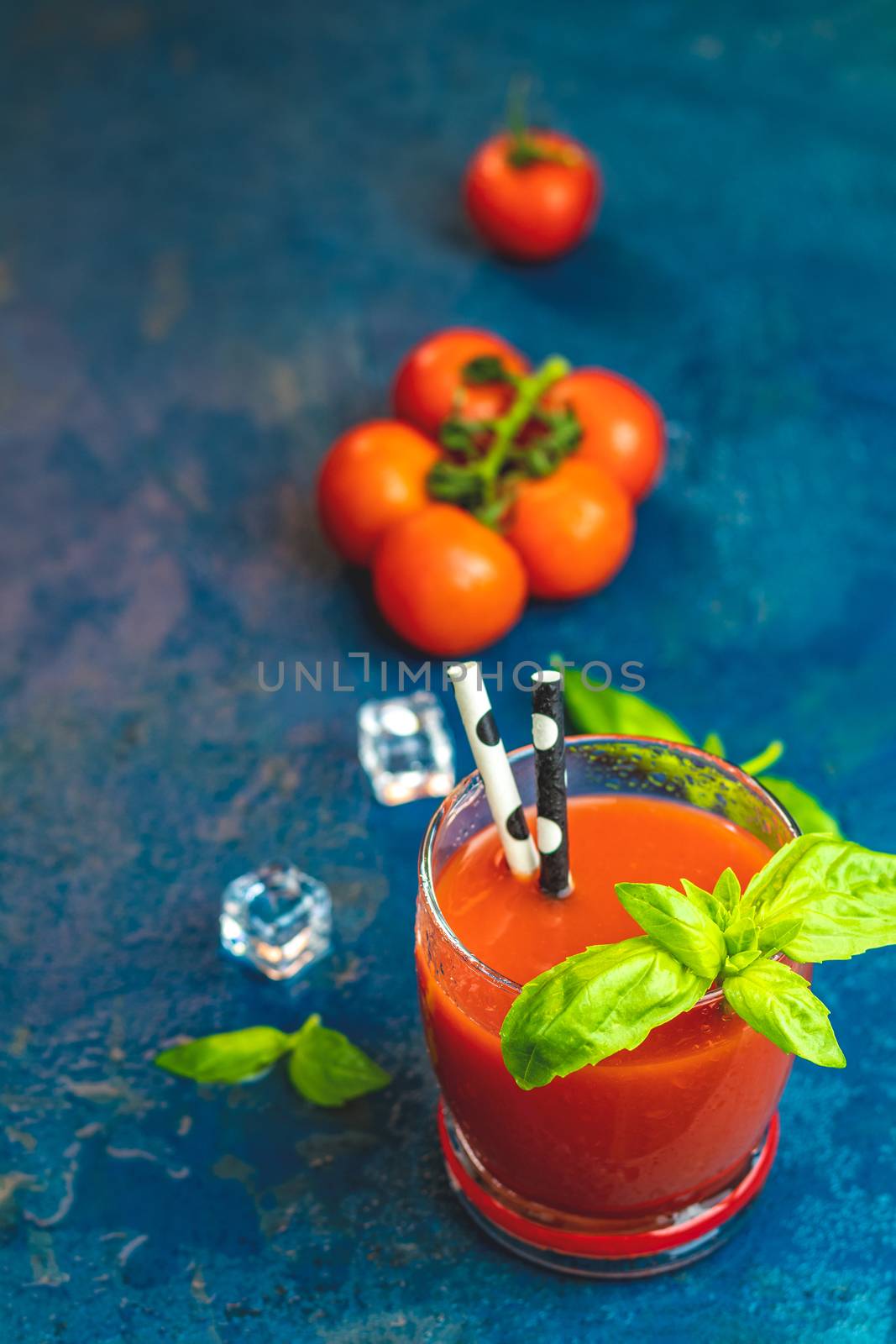 Tomato juice, red cocktail with tomato juice between tomatoes, fresh basil and ice. Delicious tomato bloody mary cocktail on dark blue concrete table.