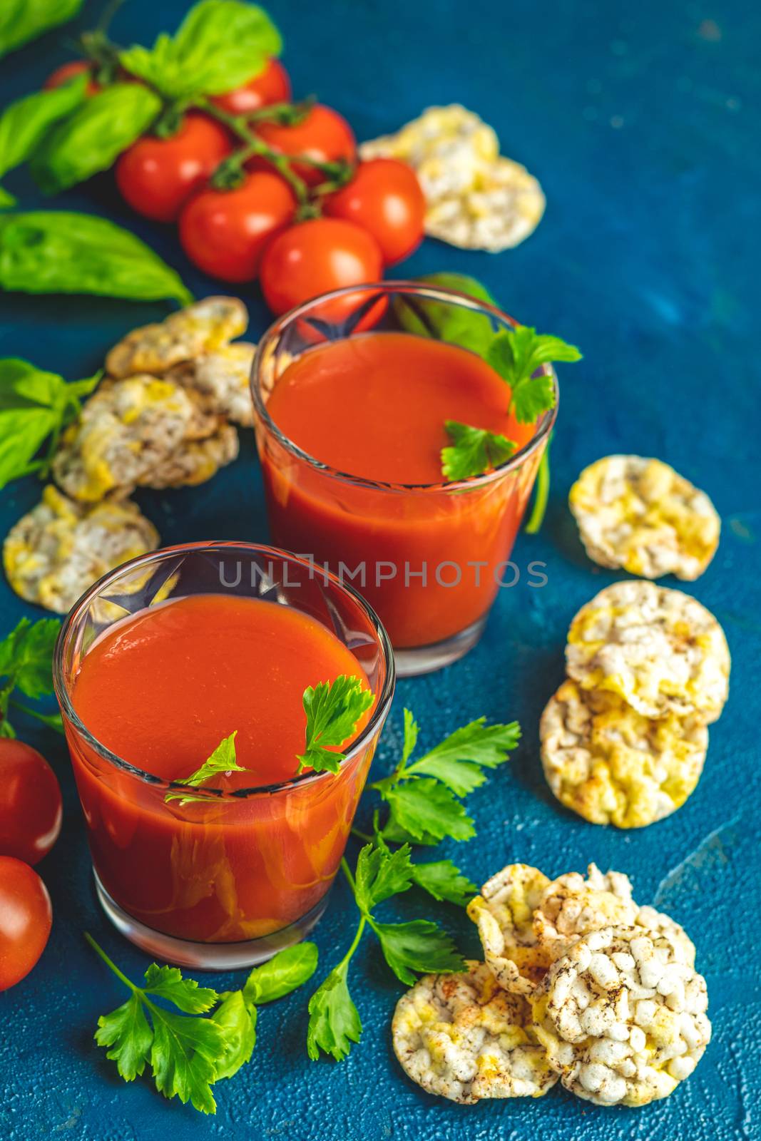 Red cocktail with tomato juice between tomatoes, basil, parsley and nutritious cereal breads. Delicious tomato bloody mary cocktail on dark blue concrete table.