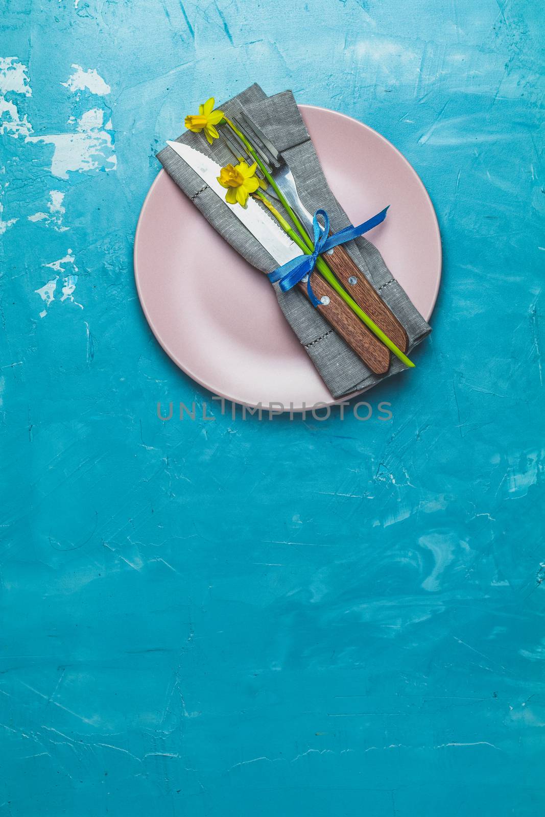 Empty pink plate and cutlery with daffodils on a napkin. Top view, green concrete surface background, copy space for you text