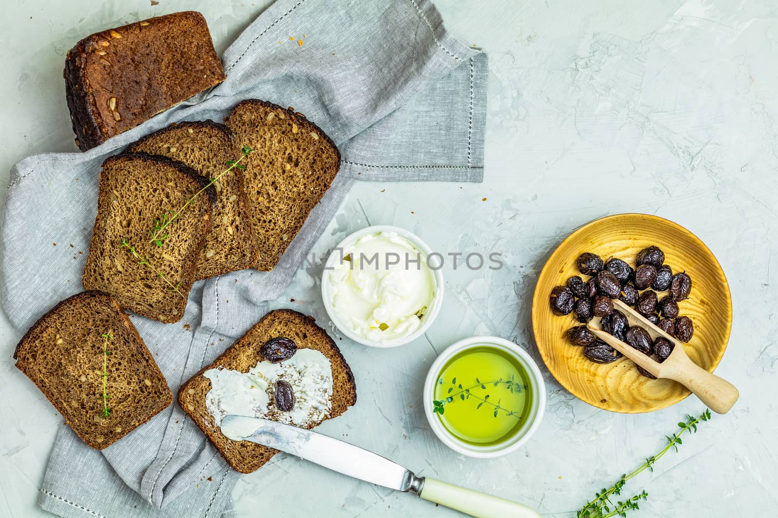 Whole wheat bread baked at home, bio ingredients, very healthy with seed, bowl of cream cheese and traditional greek italian appetizer dried olives on light gray concrete table surface.