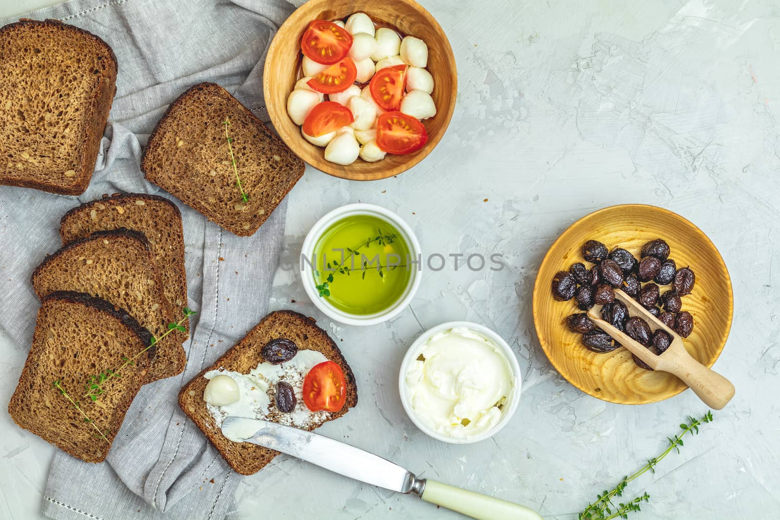 Whole wheat bread baked at home, very healthy with seed, bowl of cream cheese, mozzarella, pine nuts and traditional greek italian appetizer dried olives on light gray concrete table surface.