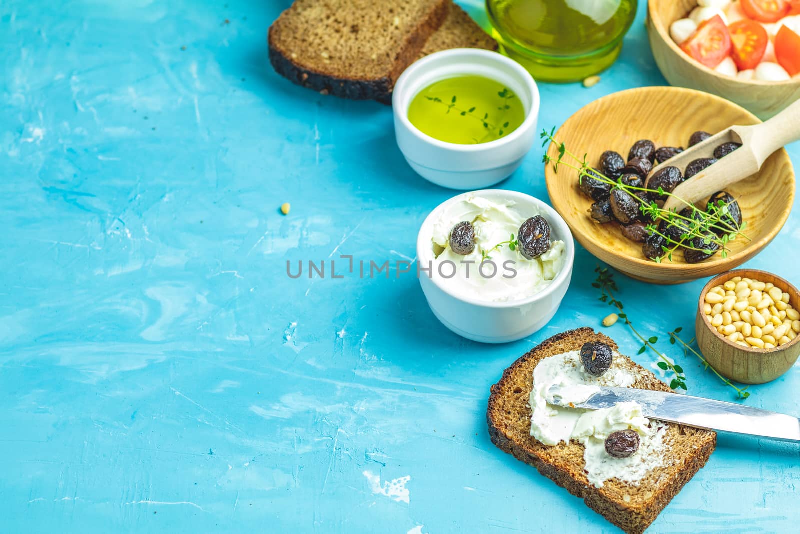 Cream Cheese on a slice of bread topped, bowl of cream cheese, mozzarella, pine nuts and traditional greek italian appetizer dried olives on blue concrete table surface.