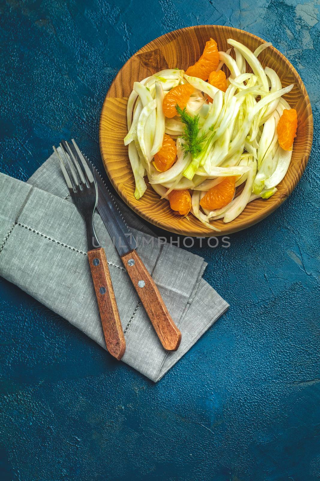 Traditional sicilian italian salad with fennel and tangerine. Fennel and orange citrus salad on wooden plate between ingredients on dark blue concrete table surface. Modern, light, raw, veg cuisine