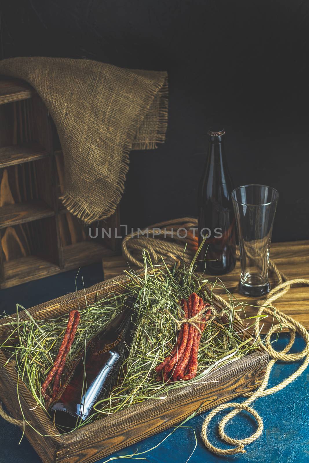 Craft beer with sausages kabanosi in the wooden box above fresh hay or dried grass beside beer bottle and drinking glass, dark rustic style.