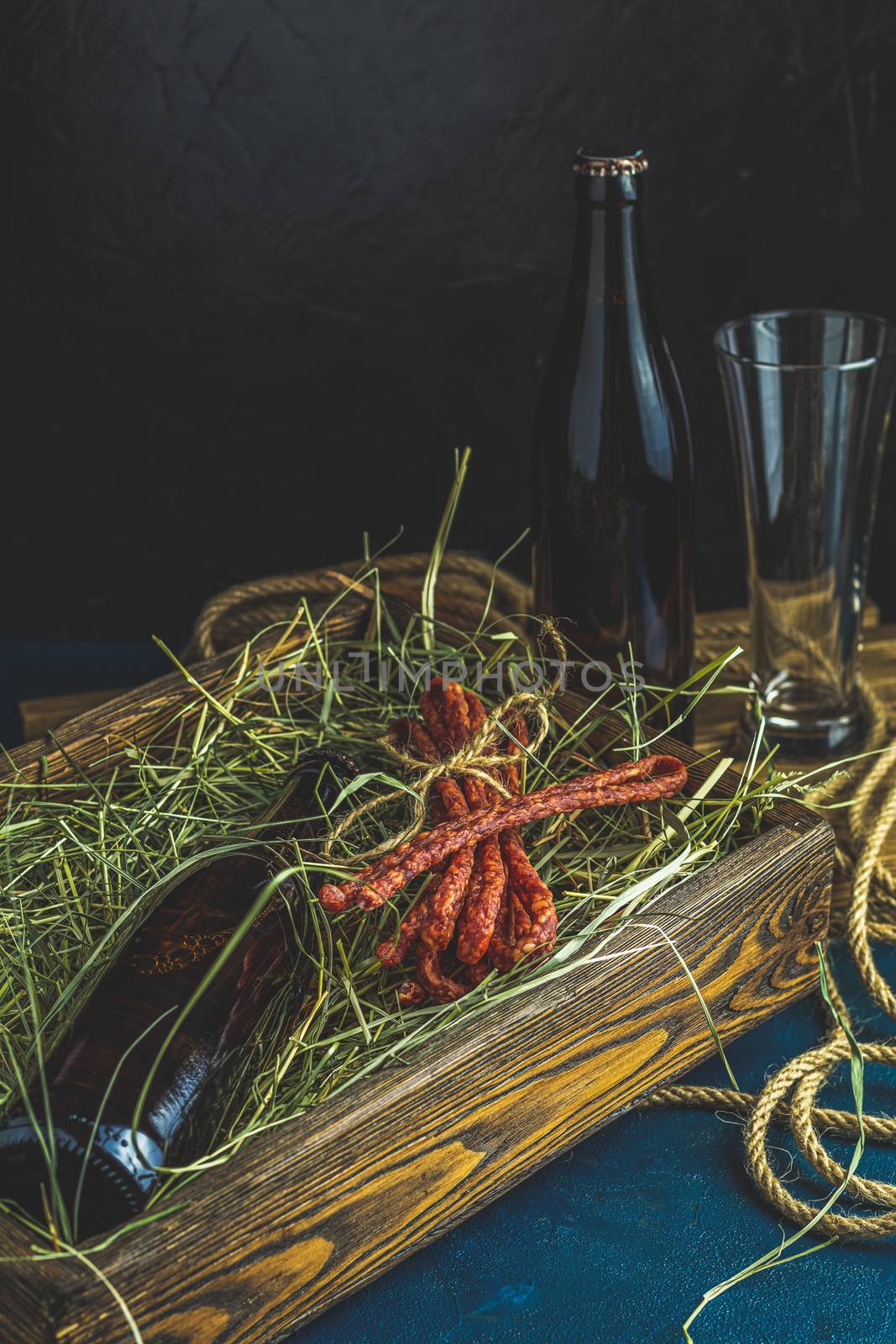 Craft beer with sausages kabanosi in the wooden box above fresh hay or dried grass beside beer bottle and drinking glass, dark rustic style.
