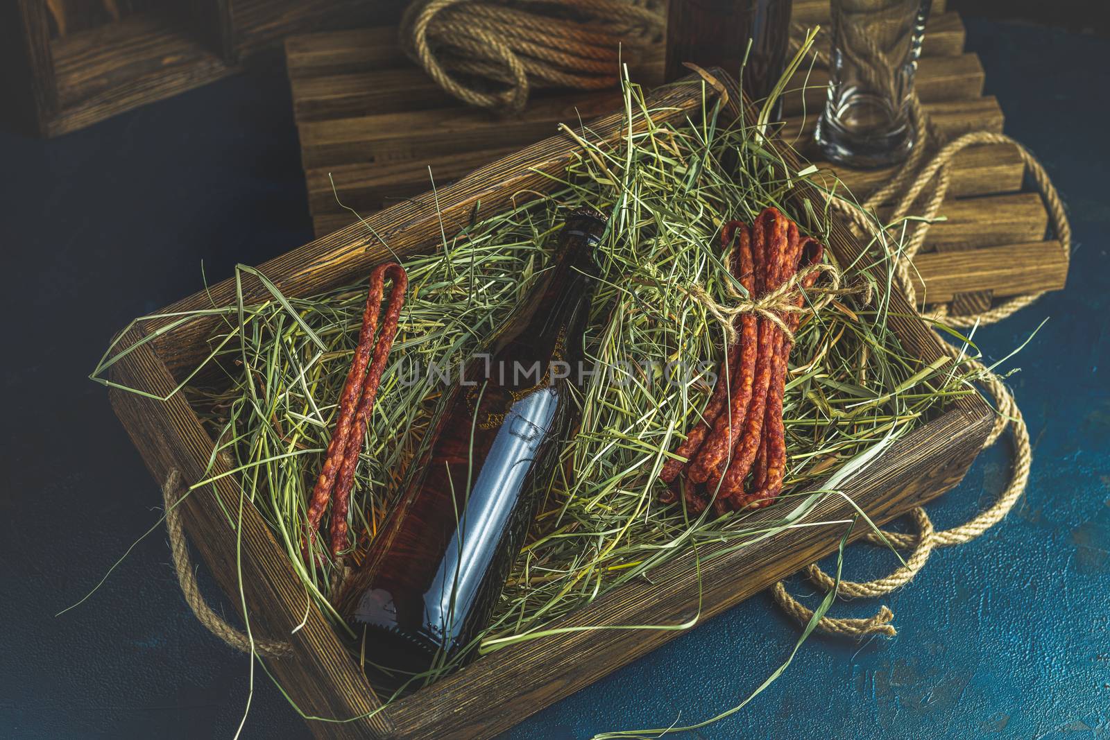 Craft beer with sausages kabanosi in the wooden box above fresh hay or dried grass beside beer bottle and drinking glass, dark rustic style.