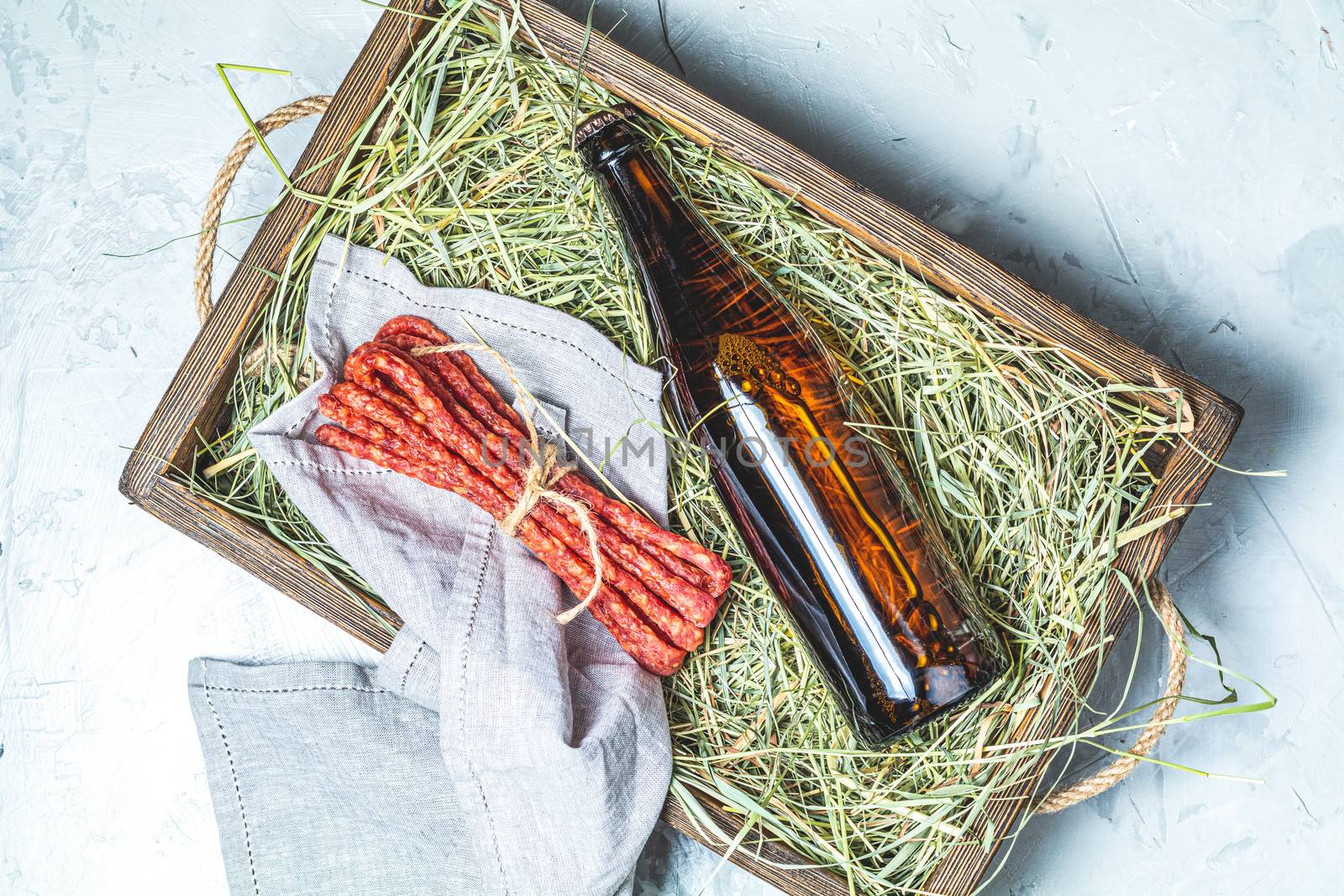 Craft beer with sausages kabanosi and dried grass in wooden box on gray concrete surface background, copy space for you text, rustic style.