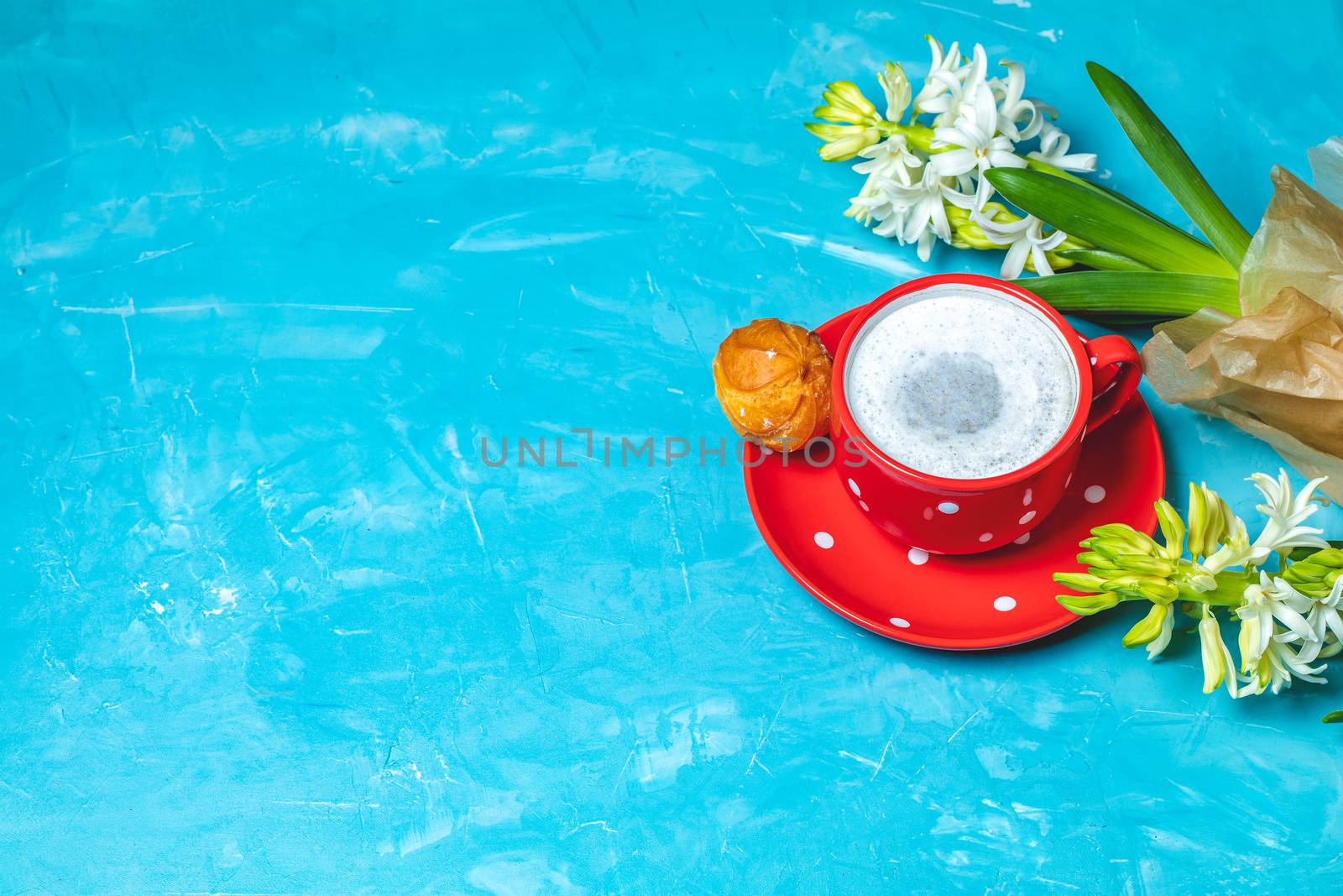 Red in white dotted cup of coffee with milk, delicious profiteroles with cream and white hyacinths on blue concrete surface background, copy space. Beautiful spring greeting card.