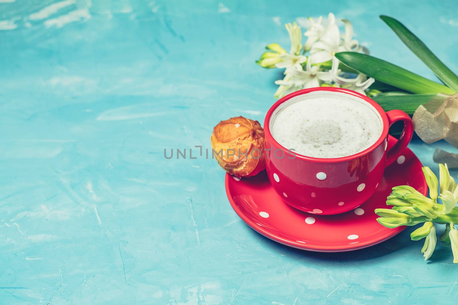Red in white dotted cup of coffee with milk, delicious profiteroles with cream and white hyacinths on blue concrete surface background, copy space. Beautiful spring greeting card.