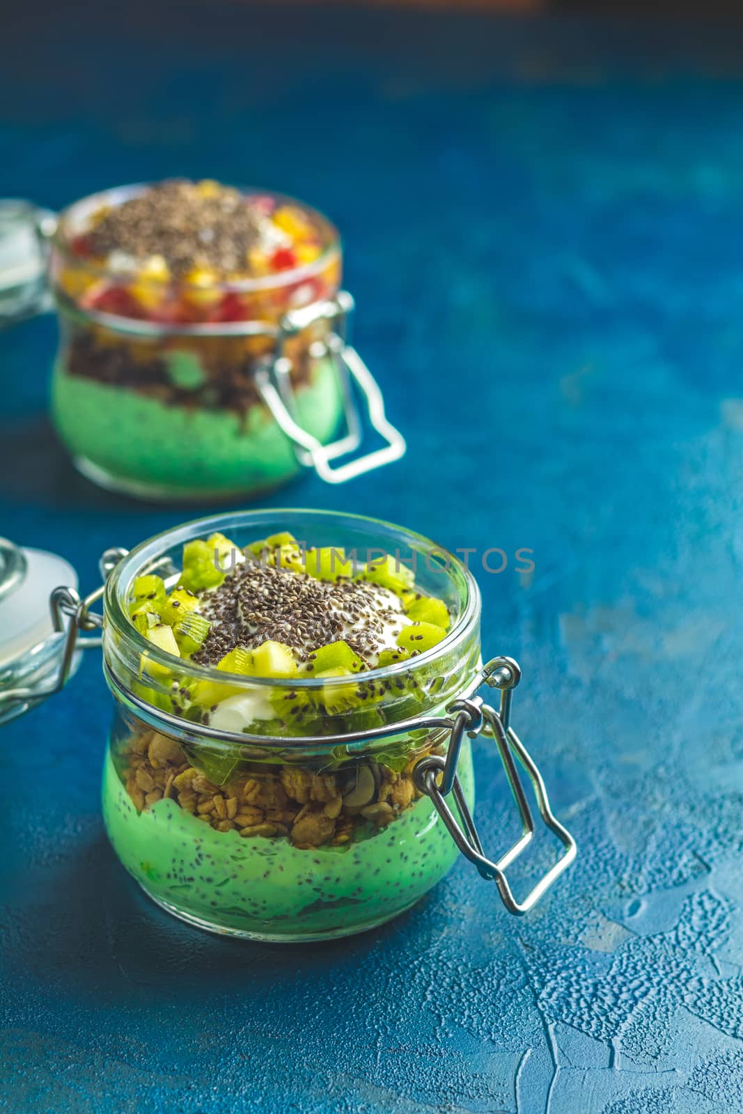 Two chia seed pudding with matcha green tea, kiwi and granola, orange in glasses on dark blue concrete background. Healthy breakfast.