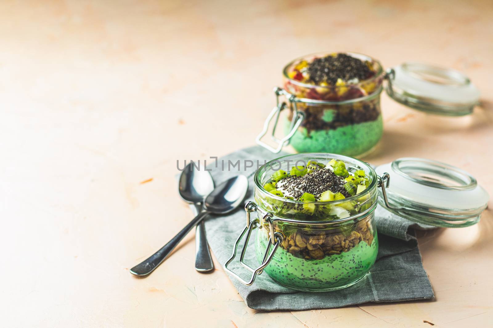 Two chia seed pudding with matcha green tea, kiwi and granola, orange in glasses on light pink concrete background. Healthy breakfast. Square image