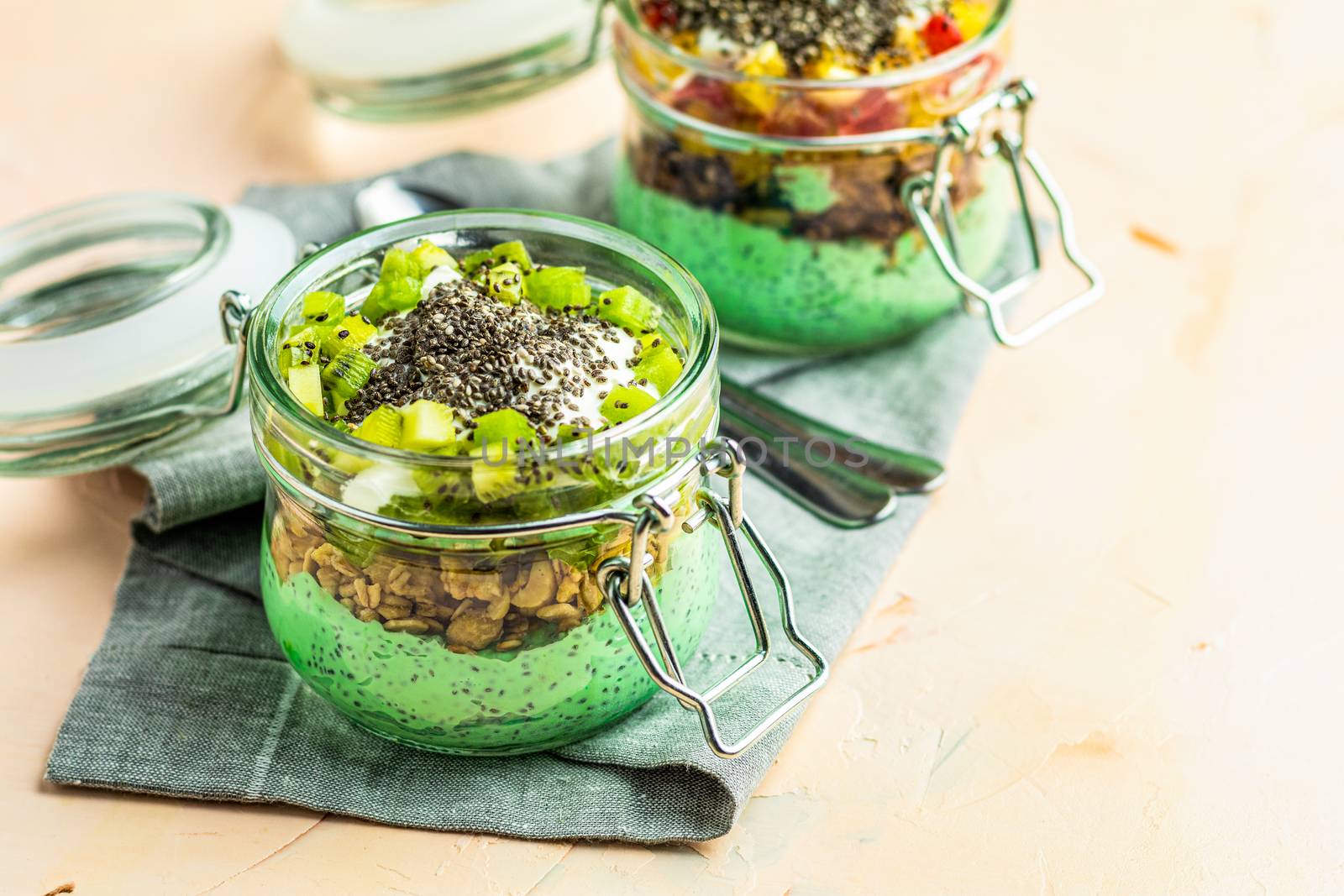 Two chia seed pudding with matcha green tea, kiwi and granola, orange in glasses on light pink concrete background. Healthy breakfast. 