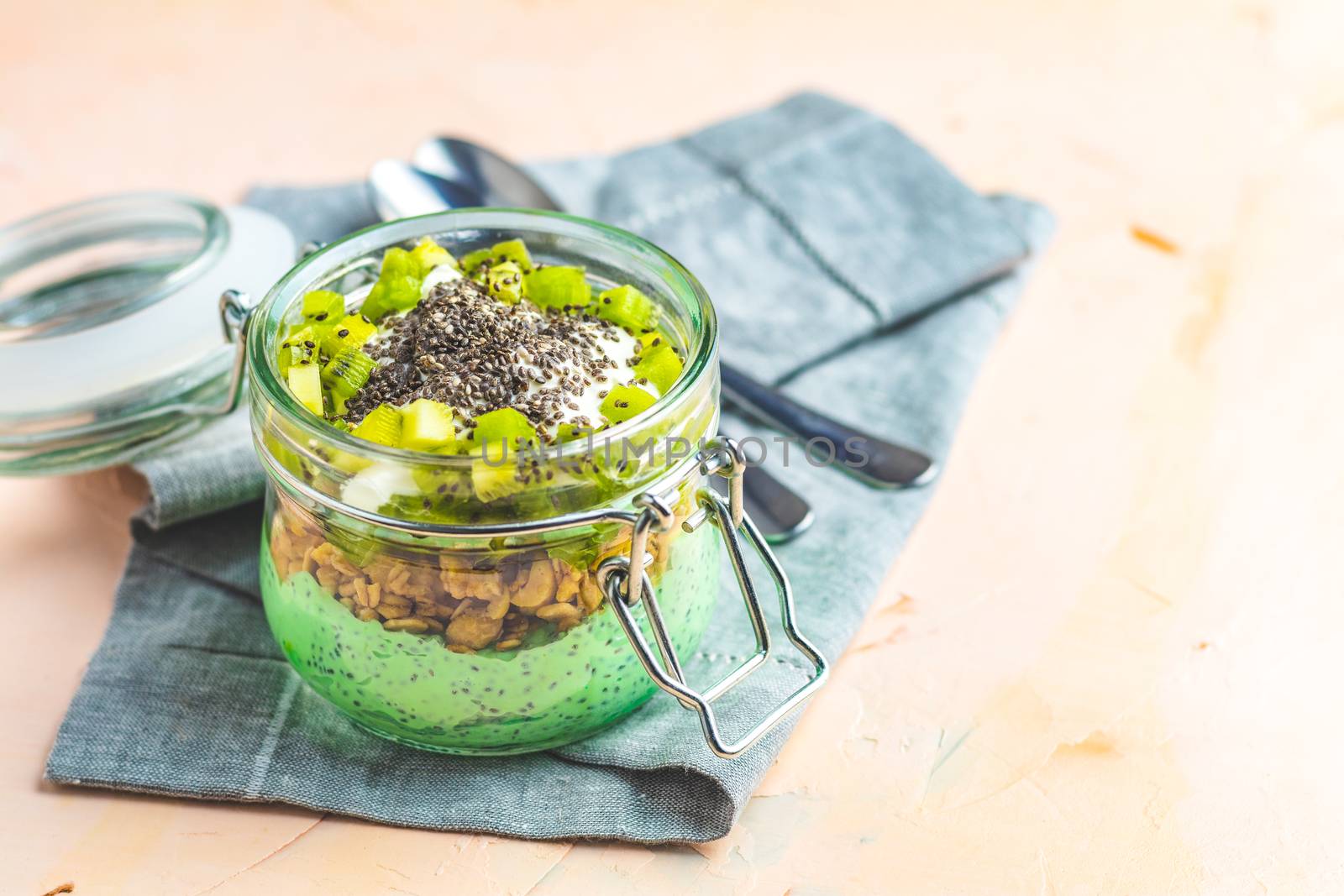 Chia seed pudding with matcha green tea, kiwi and granola in glass on light pink peachy concrete background. Healthy breakfast.