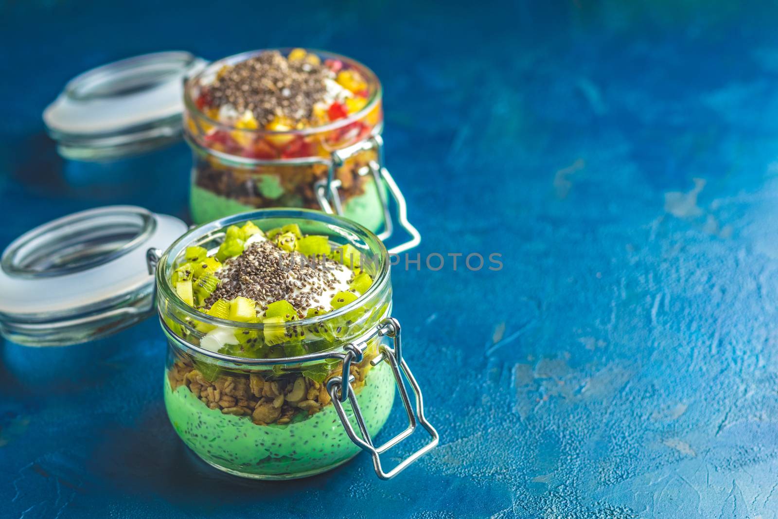 Two chia seed pudding with matcha green tea, kiwi and granola, orange in glasses on dark blue concrete background. Healthy breakfast.