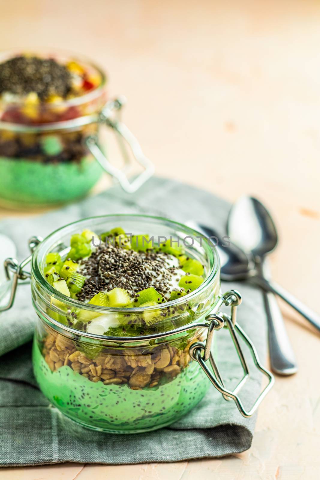 Two chia seed pudding with matcha green tea, kiwi and granola, orange in glasses on light pink concrete background. Healthy breakfast. 